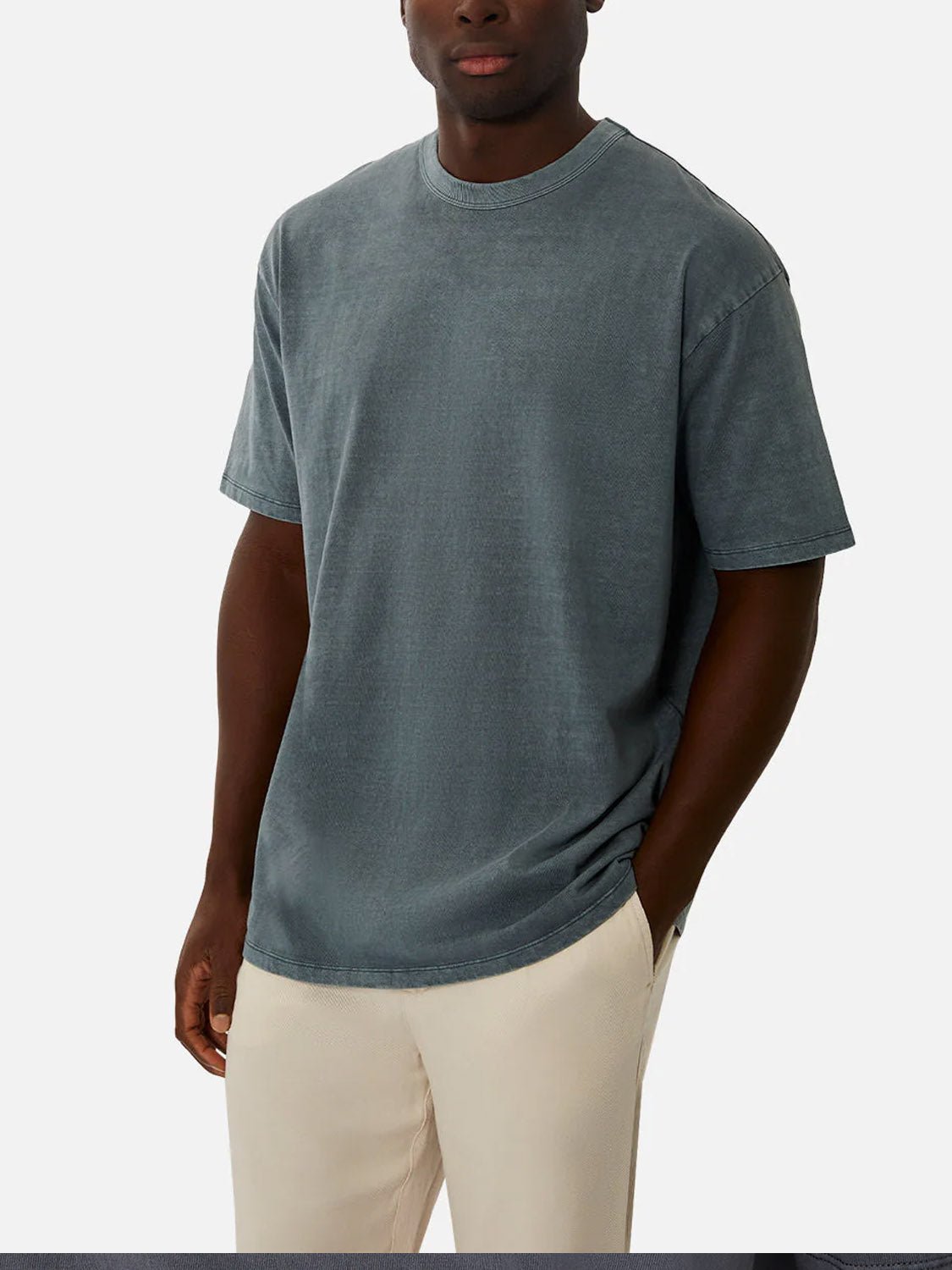 A man wearing the Industrie Del Sur Tee, a faded blue 100% cotton crew neck t-shirt, and light beige pants stands with one hand in his pocket against a plain light background.