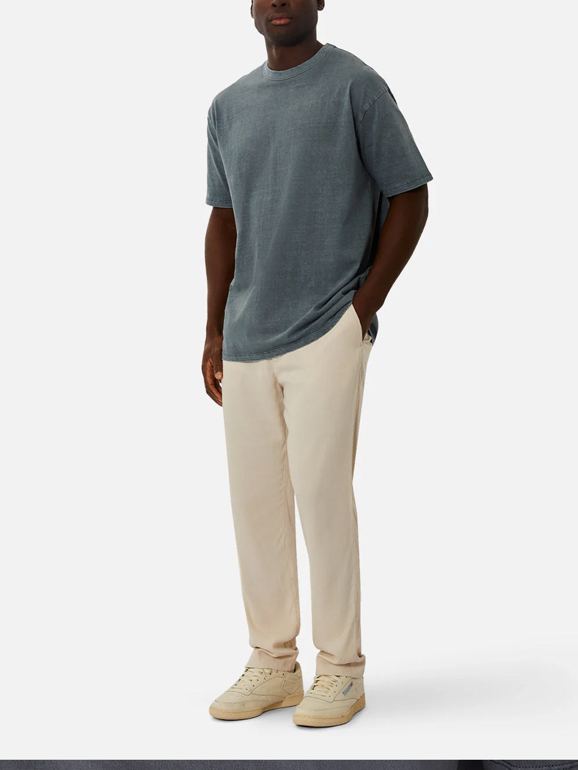 A man stands with one hand in his pocket, wearing The Del Sur Tee by Industrie—a relaxed fit, 100% cotton crew neck t-shirt in faded grey—paired with light beige pants and off-white sneakers against a plain light background.
