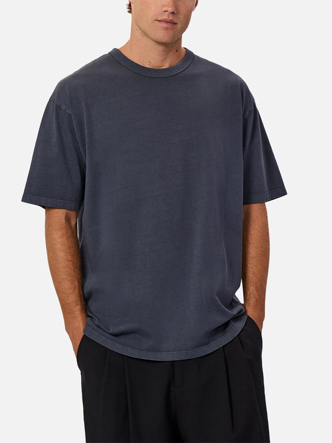 A man wearing Industrie’s The Del Sur Tee, a relaxed fit dark gray 100% cotton crew neck, pairs it with black pants and stands with his hands in his pockets against a plain light background.
