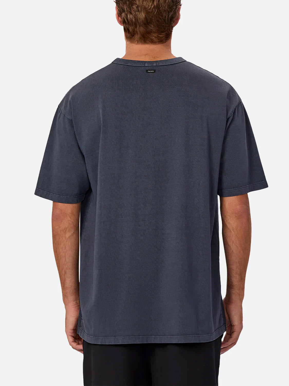 A man with short brown hair is seen from behind wearing Industrie’s The Del Sur Tee in dark gray and black shorts, standing against a plain white background.