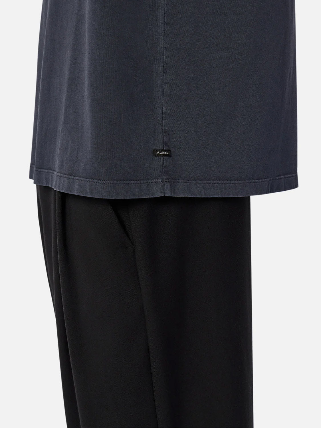 A close-up of a person wearing Industrie’s The Del Sur Tee in dark blue and black pants, with only their lower torso and upper legs visible against a plain background.