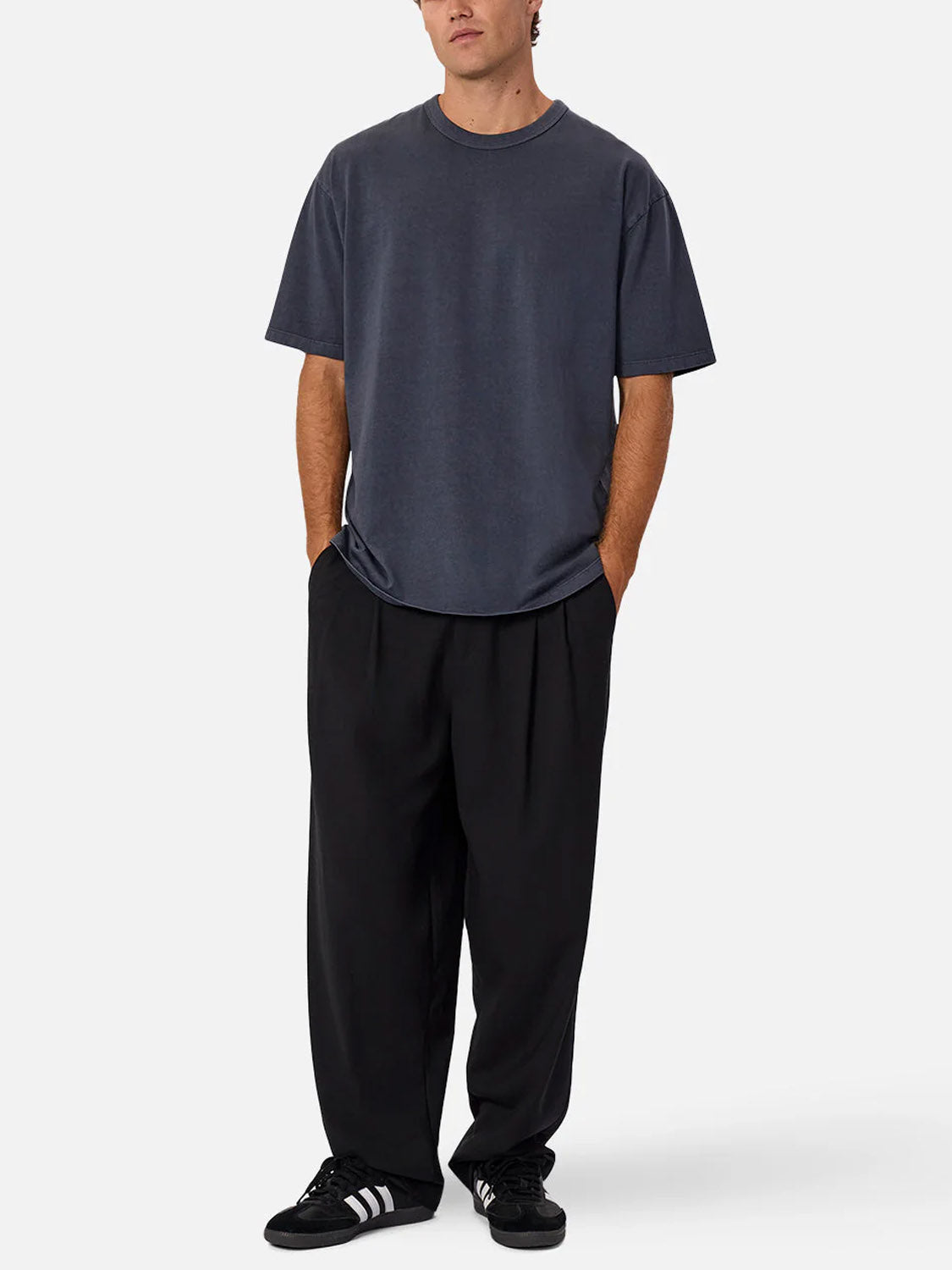 A person stands facing forward in The Del Sur Tee by Industrie, a relaxed-fit dark gray cotton crew neck, paired with black pleated wide-leg pants and black Adidas sneakers with white stripes, against a plain light background.