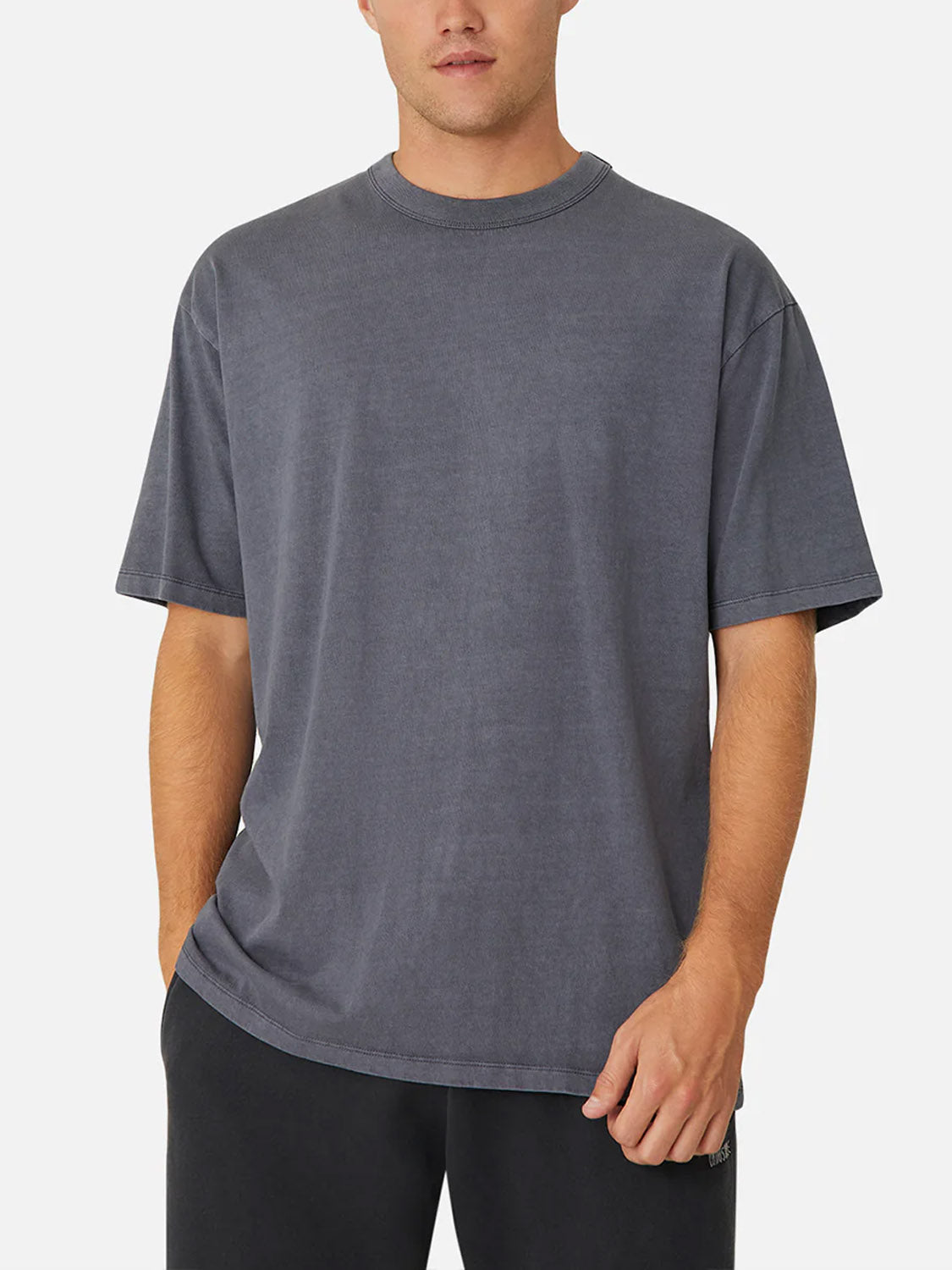 A man stands against a plain background, wearing Industrie’s The Del Sur Tee— a relaxed fit, short-sleeved dark gray 100% cotton crew neck—paired with black pants. His left hand is in his pocket and his right hand hangs by his side.