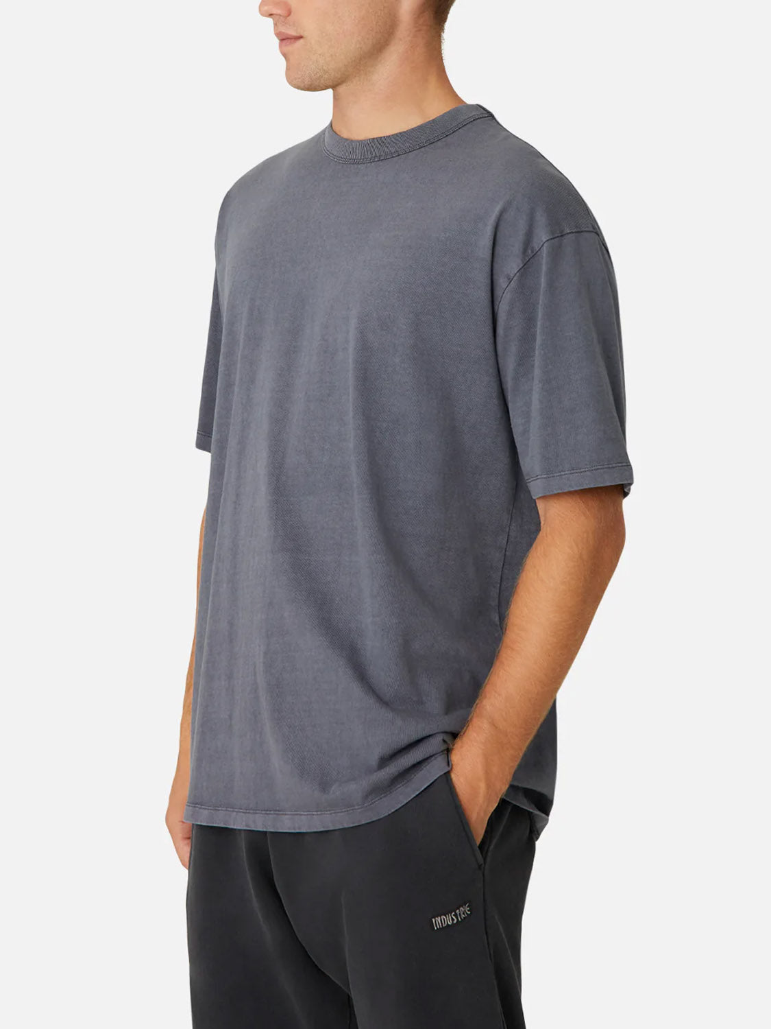A man wears the Industrie "The Del Sur Tee," a relaxed fit 100% cotton crew neck in faded gray, along with dark pants. He stands with hands in pockets, facing slightly to the side against a plain white background.