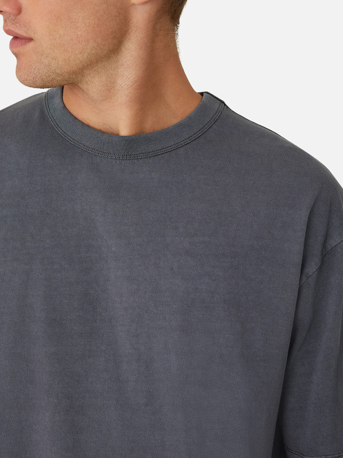 A close-up of a person wearing Industrie’s The Del Sur Tee, a relaxed fit, faded dark gray 100% cotton crew neck t-shirt, shown from the shoulders up with a plain white background and the head partially out of frame.