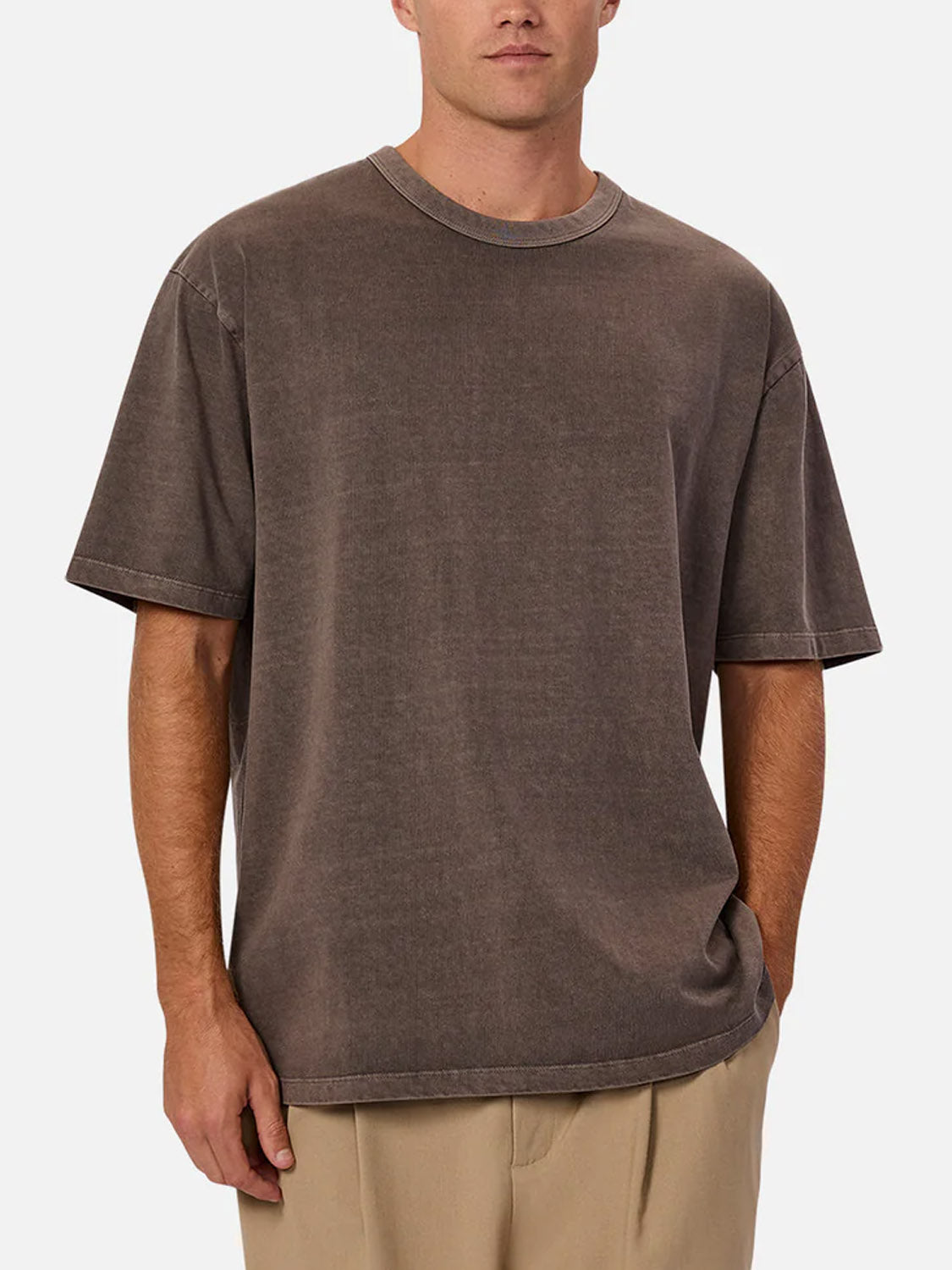 A man stands against a plain, light background wearing The Del Sur Tee by Industrie in faded brown and light beige pants, with one hand in his pocket.