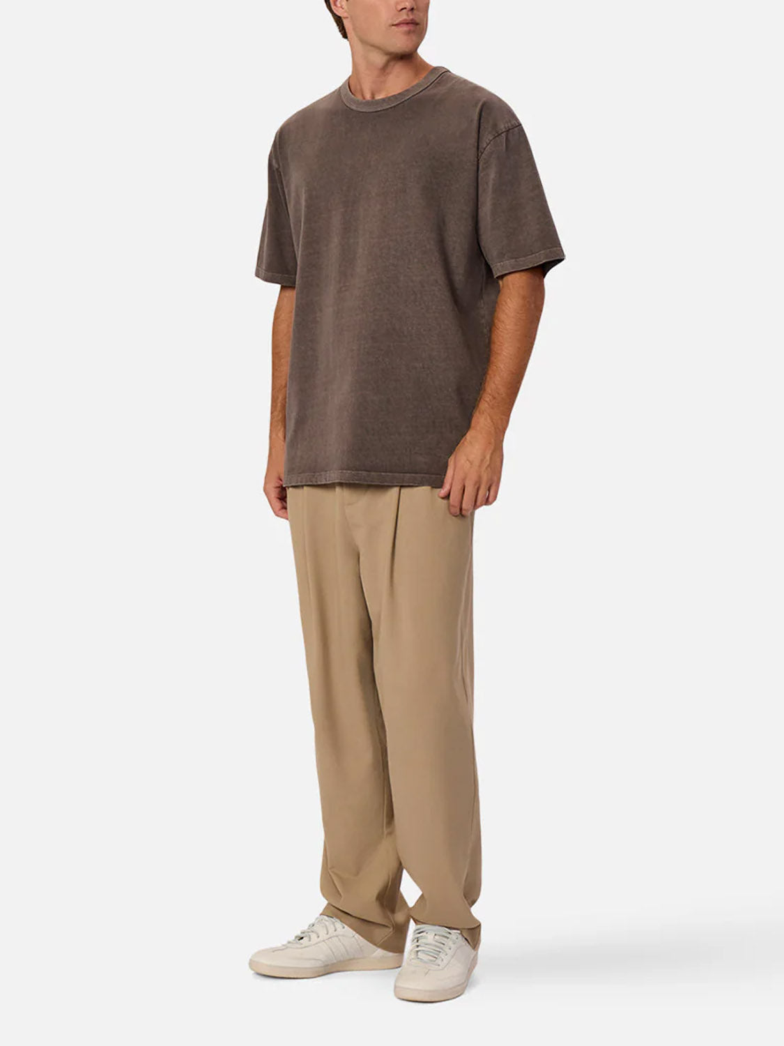 A person stands against a plain background wearing the Industrie Del Sur Tee, a relaxed fit, 100% cotton crew neck T-shirt in brown, paired with beige pleated pants and white sneakers. Their head is turned slightly, arms resting naturally by their sides.