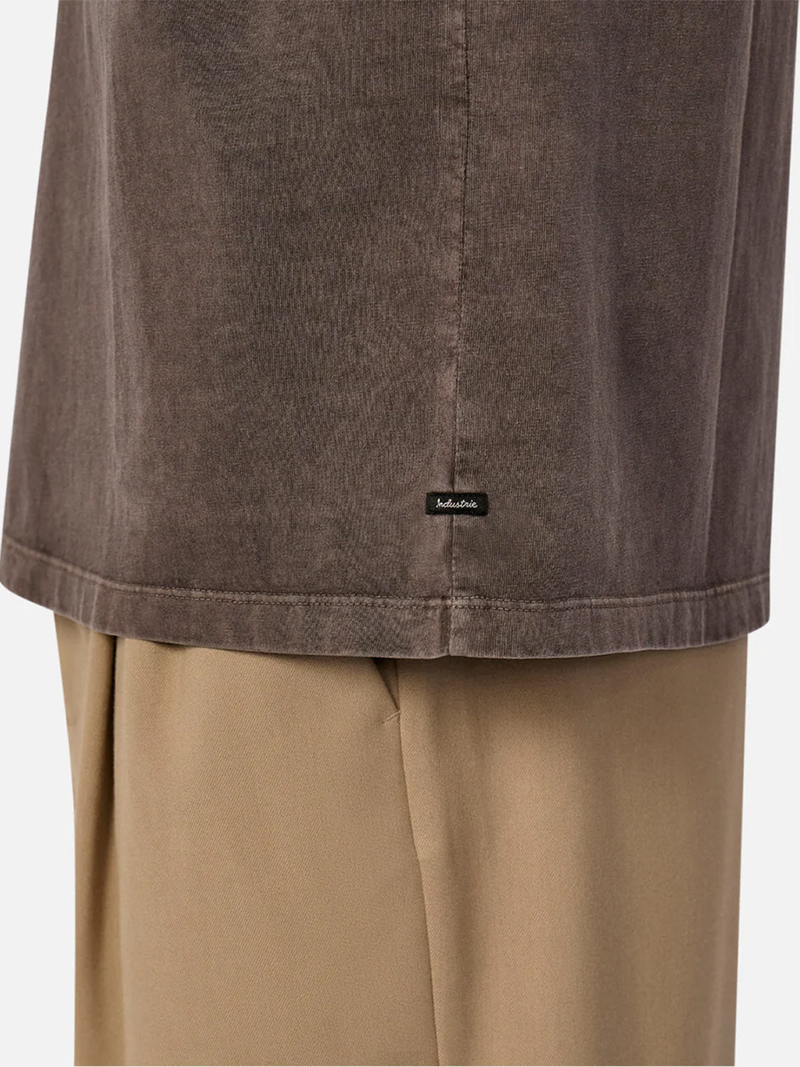A close-up of a person wearing Industrie’s The Del Sur Tee in brownish-grey over beige pants. Made from 100% cotton, the tee features a small black rectangular tag near the hem, highlighting the garments’ fabric texture.