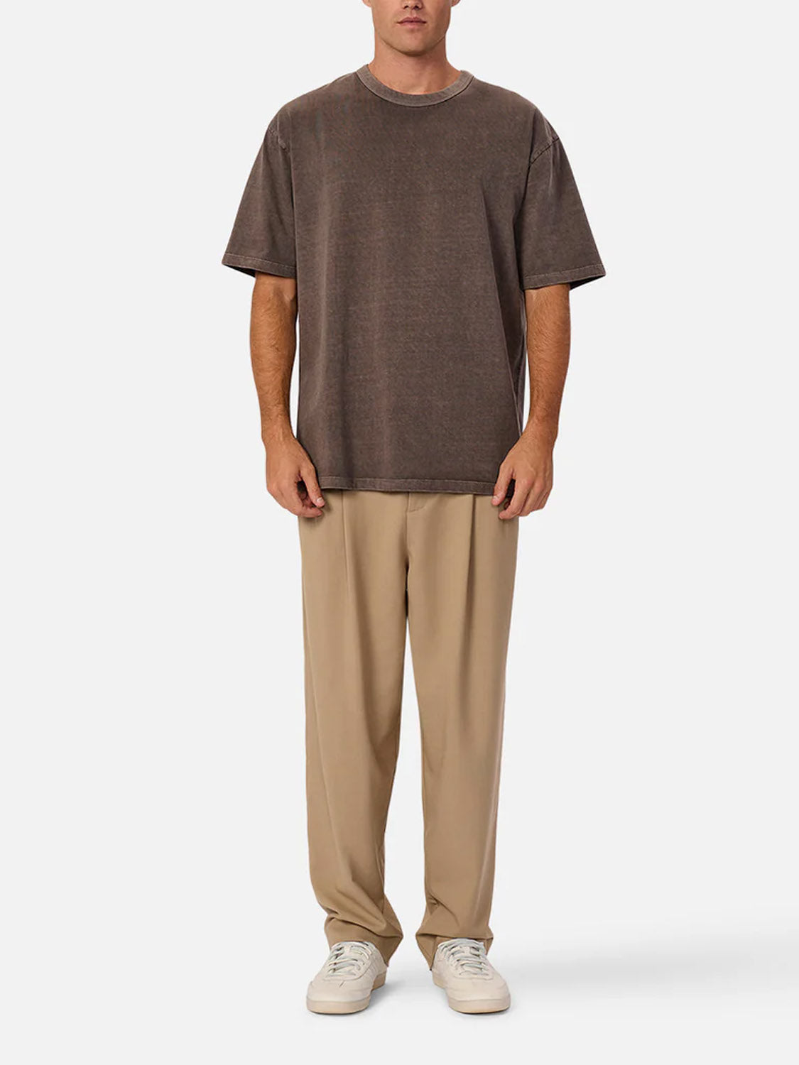 A person stands facing forward wearing Industrie’s The Del Sur Tee in dark brown, paired with light beige pants and white sneakers against a plain white background.