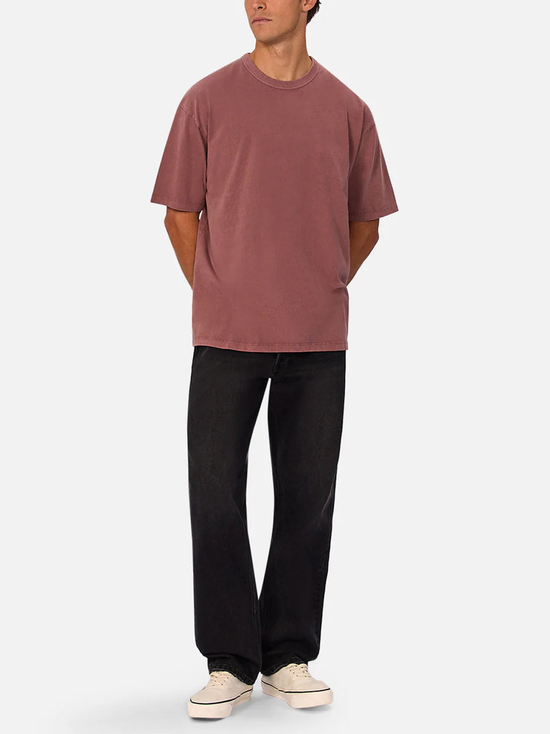 A person stands hands behind their back, wearing the Industrie Del Sur Tee—a relaxed fit, 100% cotton crew neck in mauve—styled with black jeans and white sneakers against a plain light background.