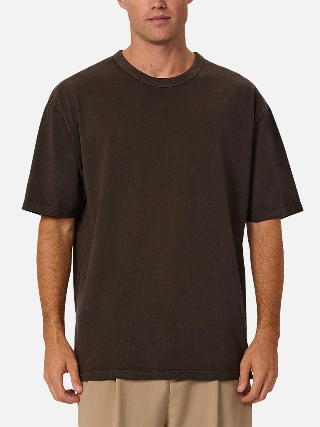 A man stands against a white background, wearing Industrie’s The Del Sur Tee in plain dark brown, relaxed fit, 100% cotton, paired with light beige pants. His face is partially visible and his arms rest at his sides.