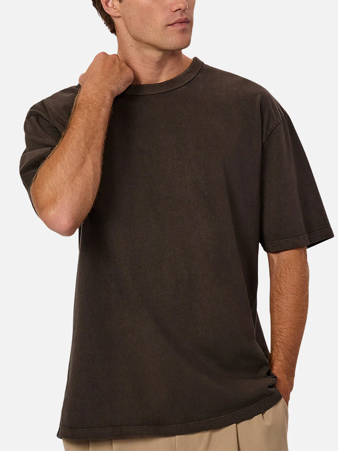 A man stands against a plain background, one hand on his neck and the other in his pocket, wearing Industrie’s The Del Sur Tee in relaxed fit dark brown 100% cotton with beige pants. His face is partially visible.