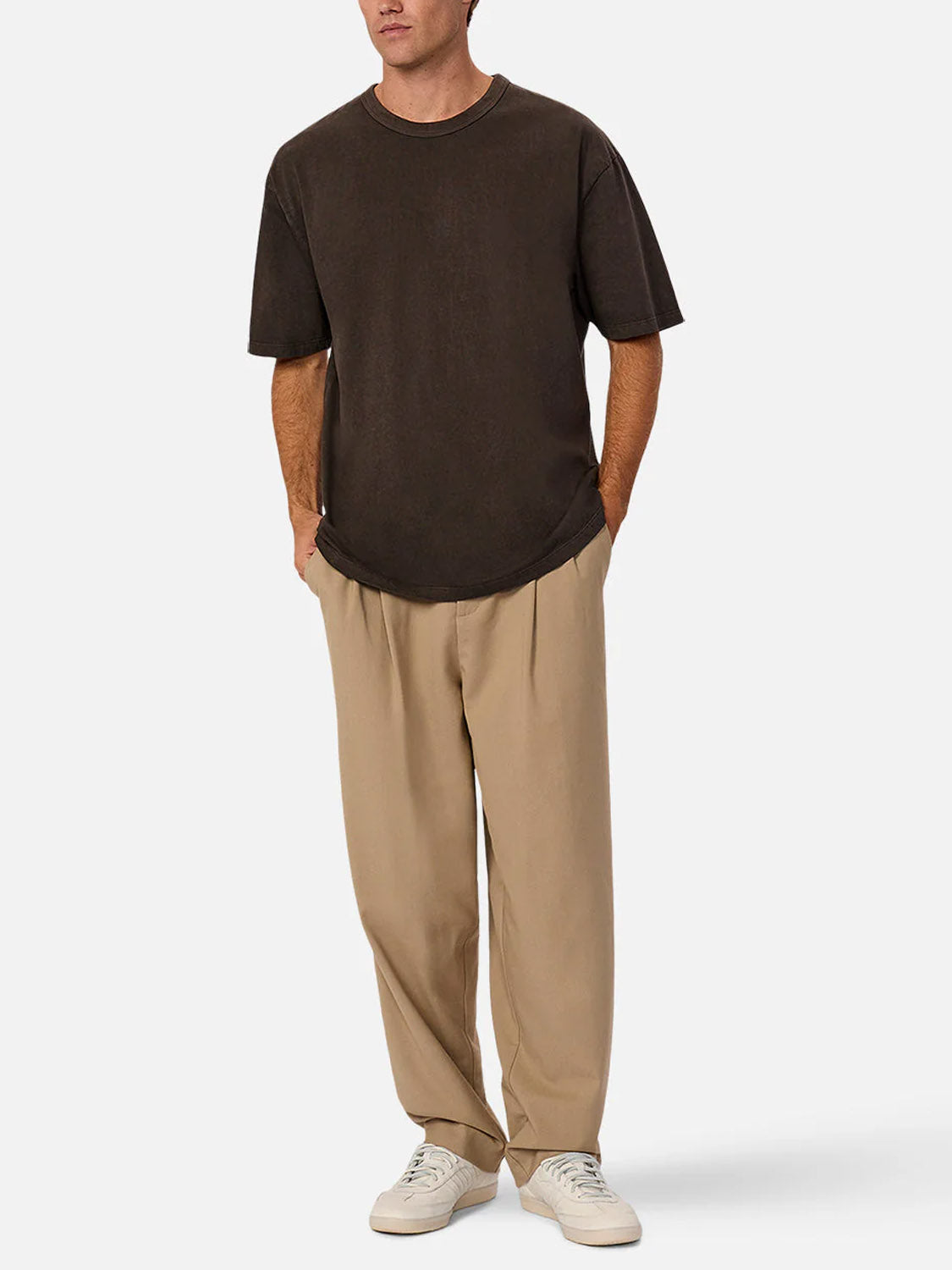 A man stands against a plain white background with his hands in his pockets, wearing the Industrie Del Sur Tee in dark brown, paired with beige pleated trousers and white sneakers.