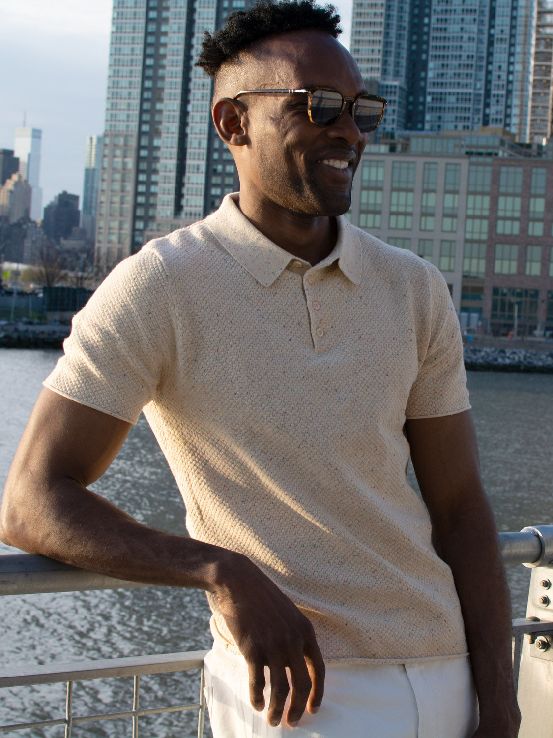 A man wearing sunglasses, white pants, and the Denver Speckled Knit Cotton Polo from ID Menswear stands by a waterfront railing, smiling with tall city buildings in the background.