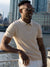 A man wearing sunglasses, white pants, and the Denver Speckled Knit Cotton Polo from ID Menswear stands by a waterfront railing, smiling with tall city buildings in the background.