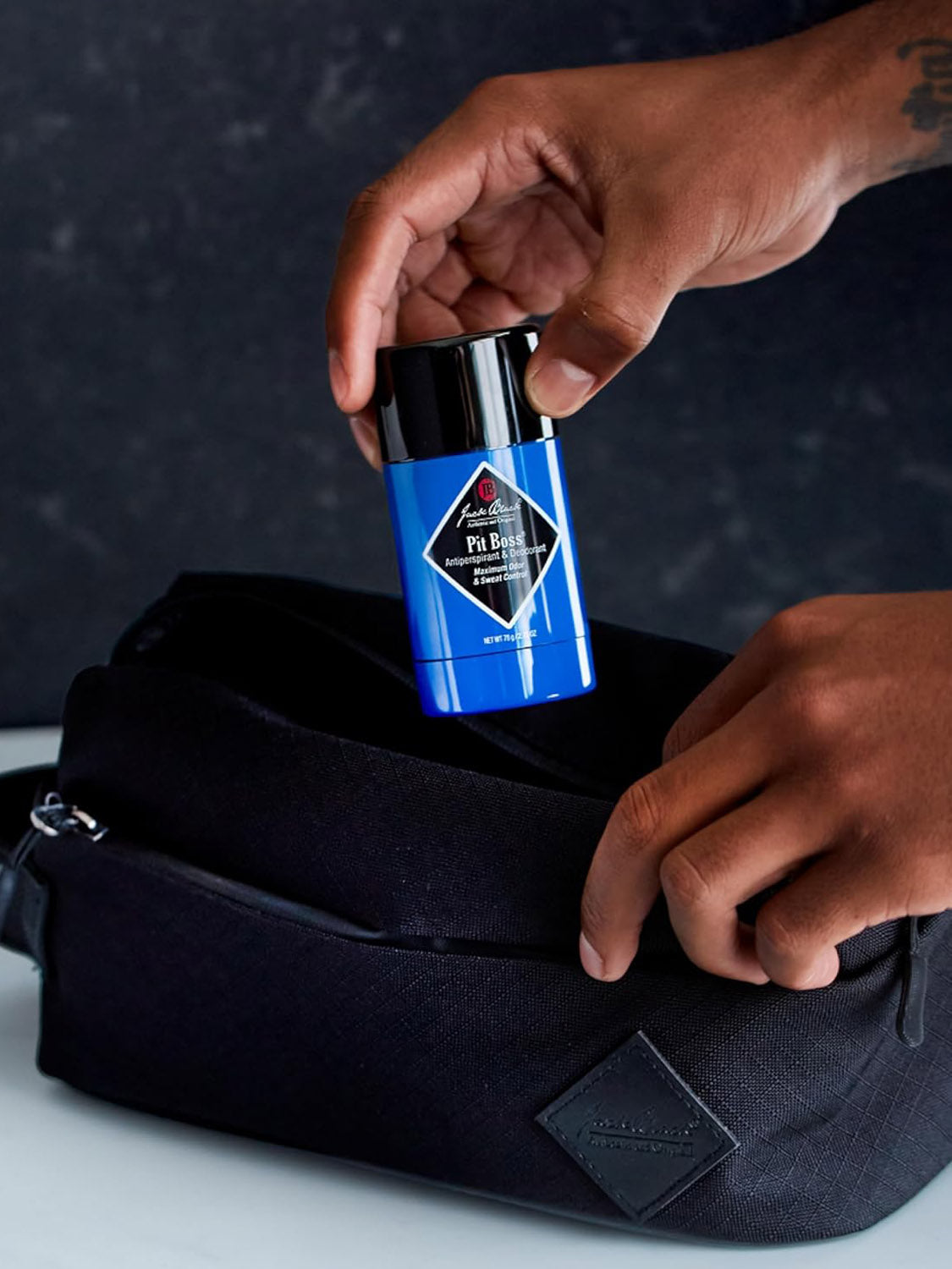 A person puts a stick of Jack Black Original Pit Boss® Antiperspirant & Deodorant into a black toiletry bag against a dark background.