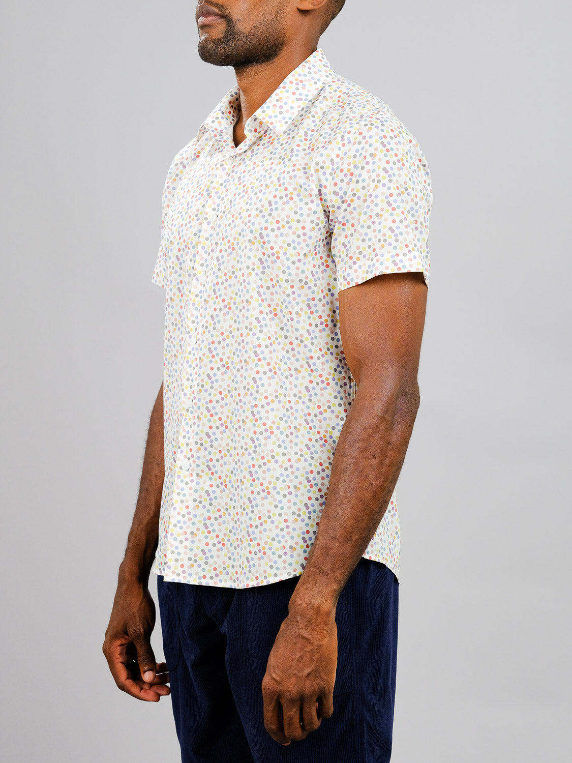 A man in the Disco Printed Cotton Shirt by ID Menswear—lightweight, short-sleeved, with vibrant multicolored polka dots—pairs it with dark pants as he stands sideways against a plain gray background.