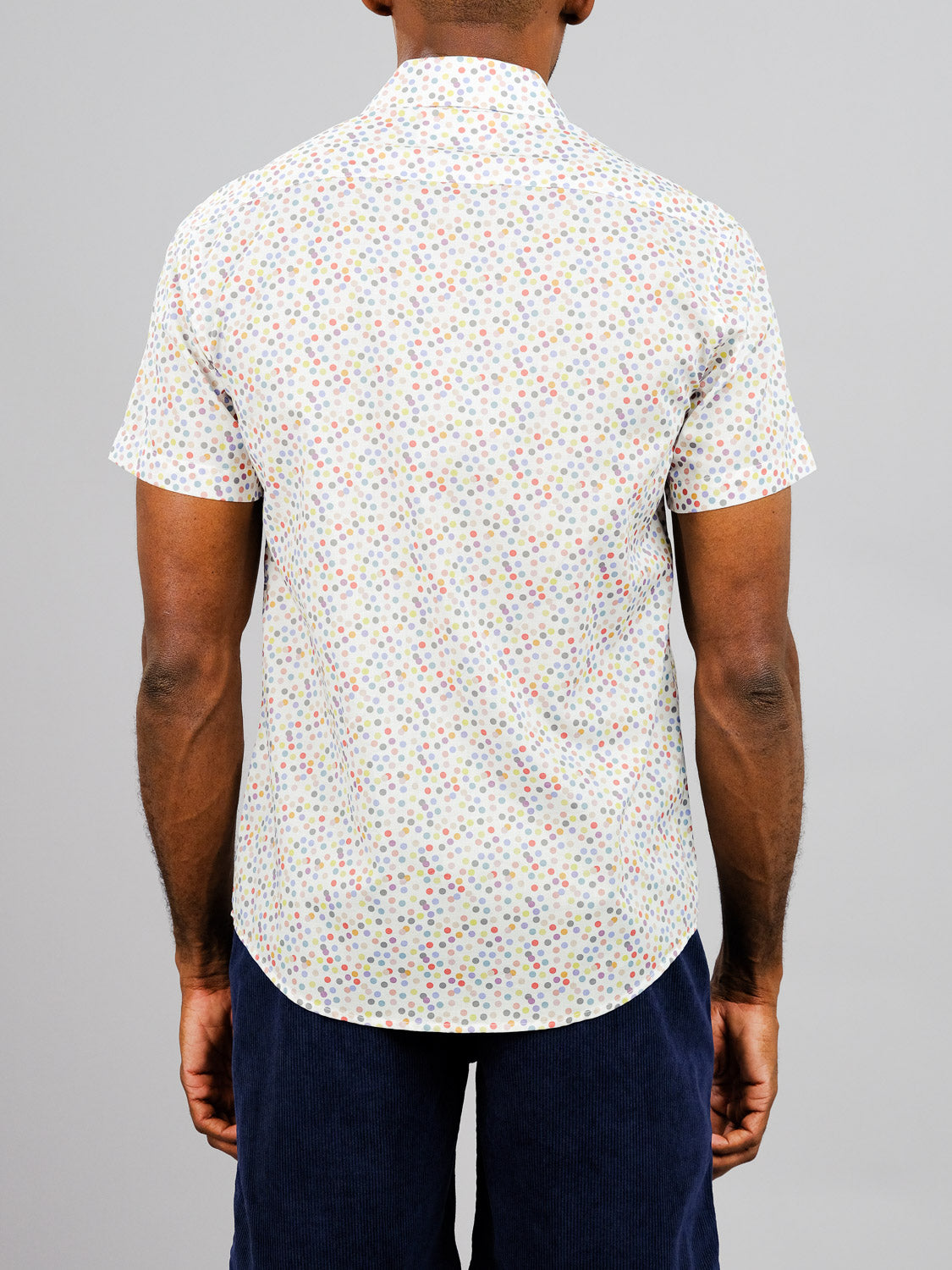 A person is seen from behind wearing the ID Menswear Disco Printed Cotton Shirt, a lightweight short-sleeved cotton shirt with a colorful polka dot pattern, paired with dark blue corduroy pants against a plain gray background.