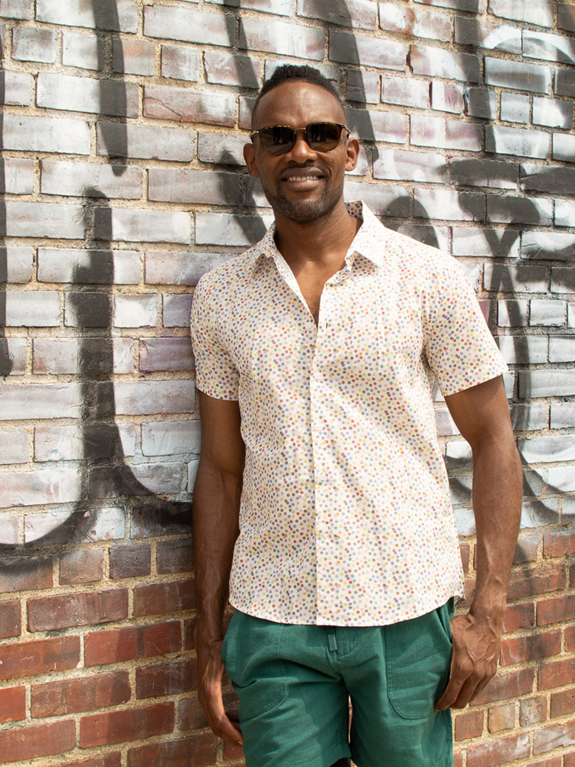 A man smiles against a graffiti-covered brick wall, wearing the Disco Printed Cotton Shirt by ID Menswear, sunglasses, and green pants.