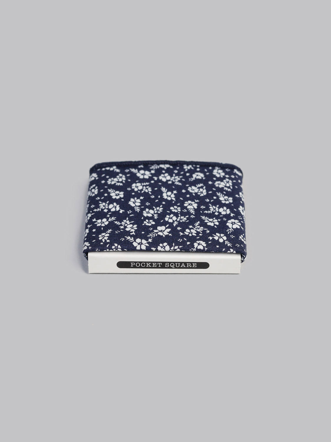 The Ditsy Pocket Square by ID Menswear is a navy blue, 100% cotton pocket square with a white floral pattern, handmade in Brooklyn. It comes neatly packaged and displayed against a plain grey background.