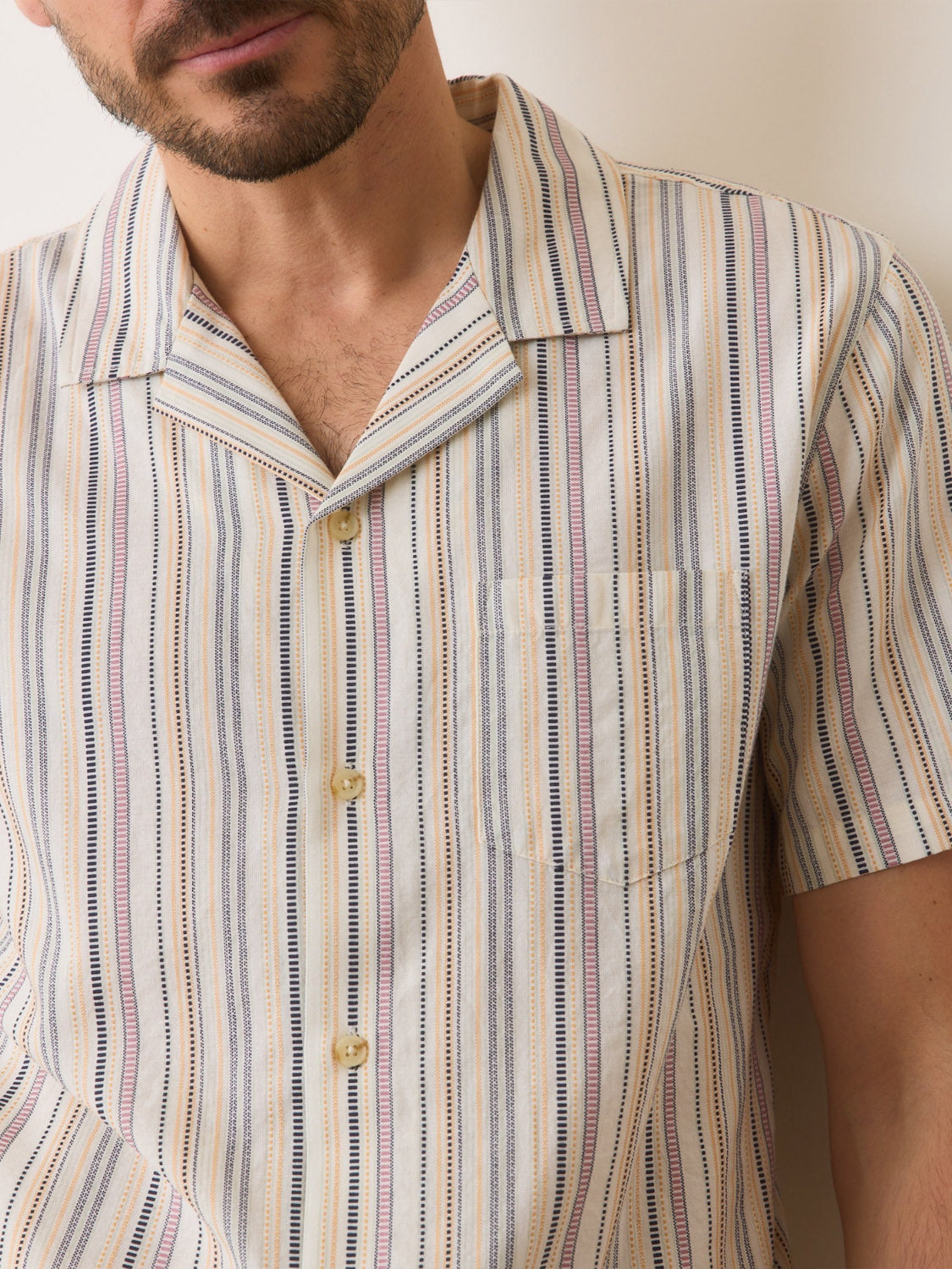 A man wears the Marine Layer Dobby Resort Shirt, a short-sleeved, button-up shirt featuring multi-colored woven stripes. Its crafted from lightweight, breathable fabric and includes a small chest pocket, set against a light-colored background. Brand: Marine Layer.