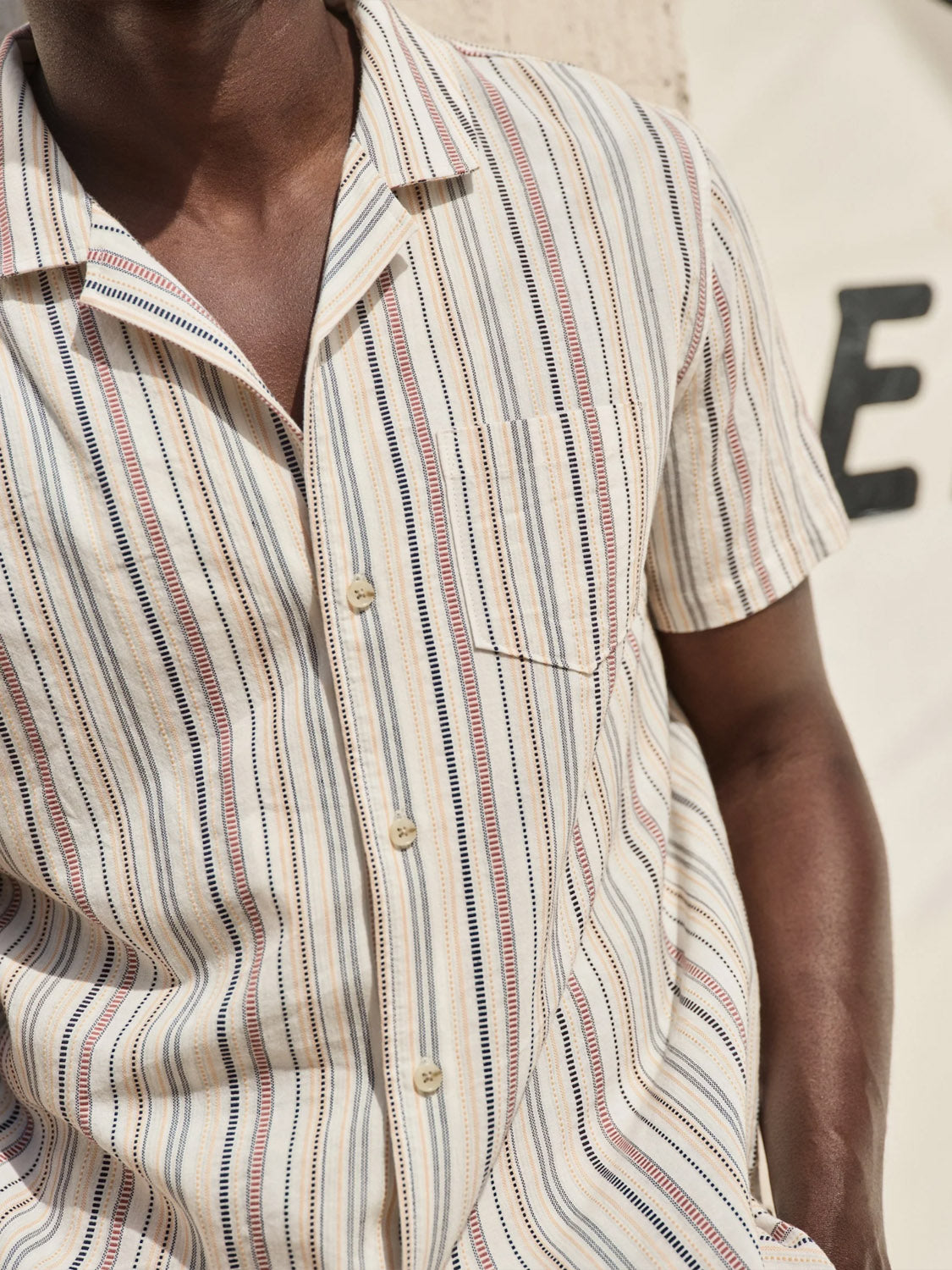Dressed in a Marine Layer Dobby Resort Shirt, their look includes the vintage charm of short sleeves and woven stripes in beige, blue, and red. The shirt features lightweight fabric with a chest pocket. A partial letter E is visible in the backdrop.