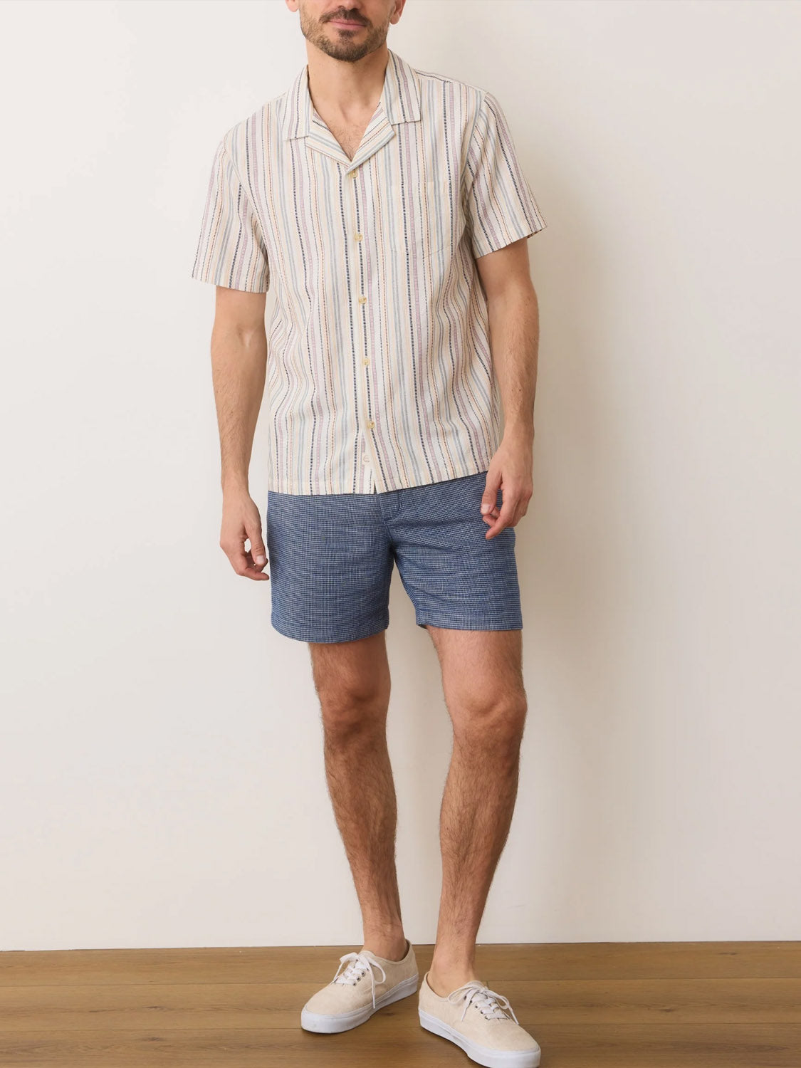 A person in a Marine Layer Dobby Resort Shirt with woven stripes and blue shorts stands on a wooden floor against a plain white wall. The outfit is completed with white sneakers, ideal for keeping cool in breathable, lightweight fabric.