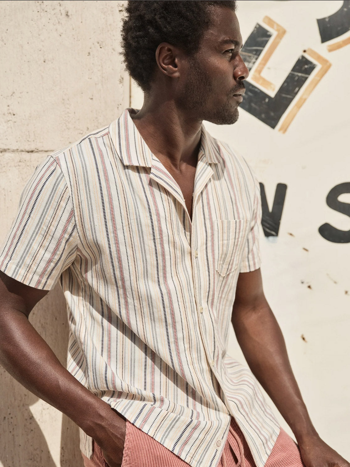 A man wearing a Marine Layer Dobby Resort Shirt and patterned shorts leans against a concrete wall with a partially visible sign in the background.