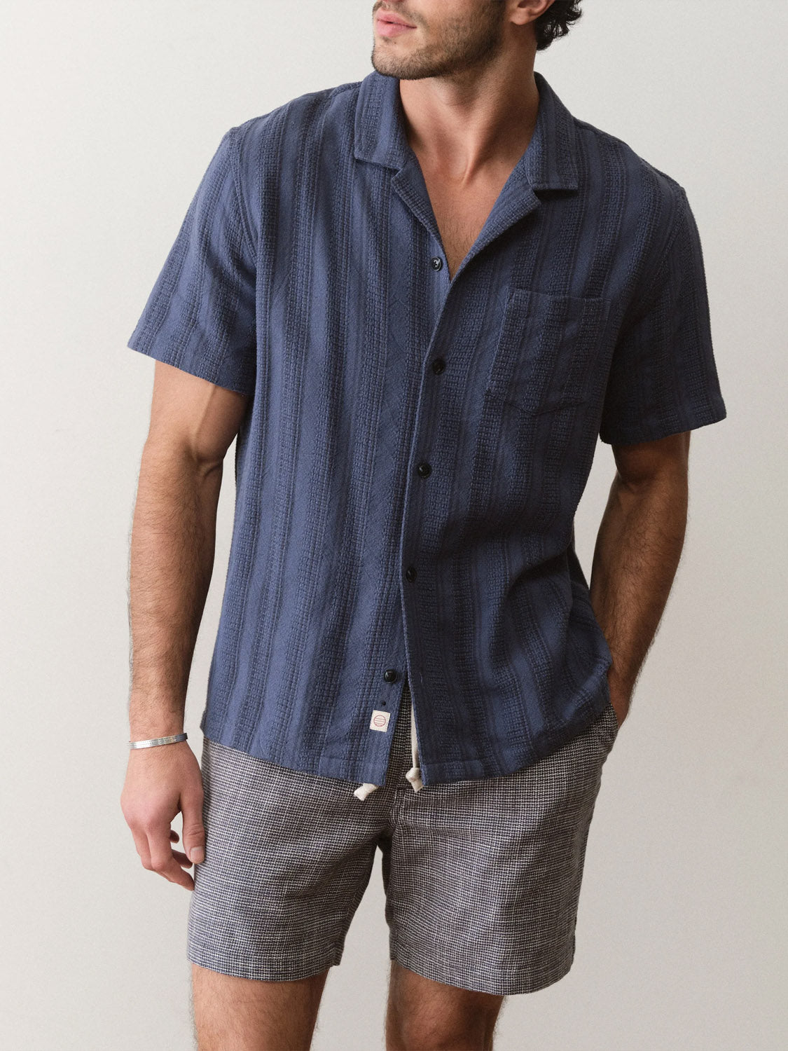 A man wears the Marine Layer Dobby Resort Shirt, a short-sleeve dark blue textured cotton piece, with gray shorts. He stands against a plain light background, head slightly turned, and one hand in his pocket.