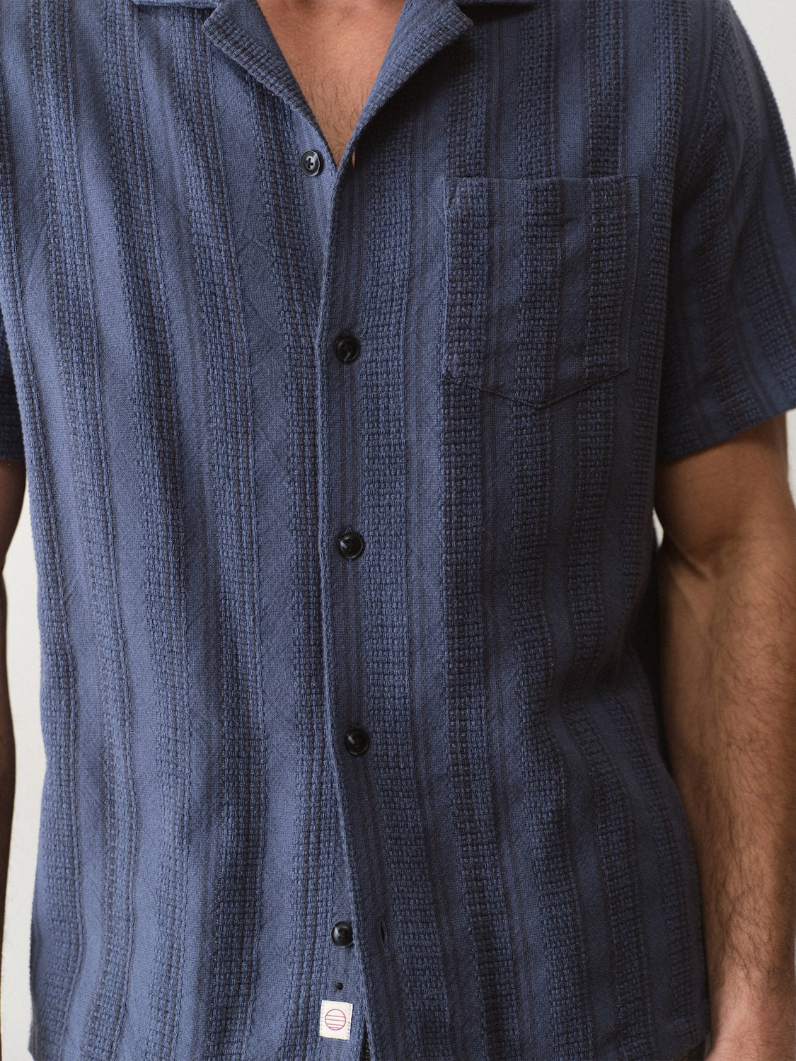 A person wears the Marine Layer Dobby Resort Shirt, a navy blue, short-sleeve, textured cotton shirt with black buttons and a chest pocket; only their torso and part of their arms are visible.