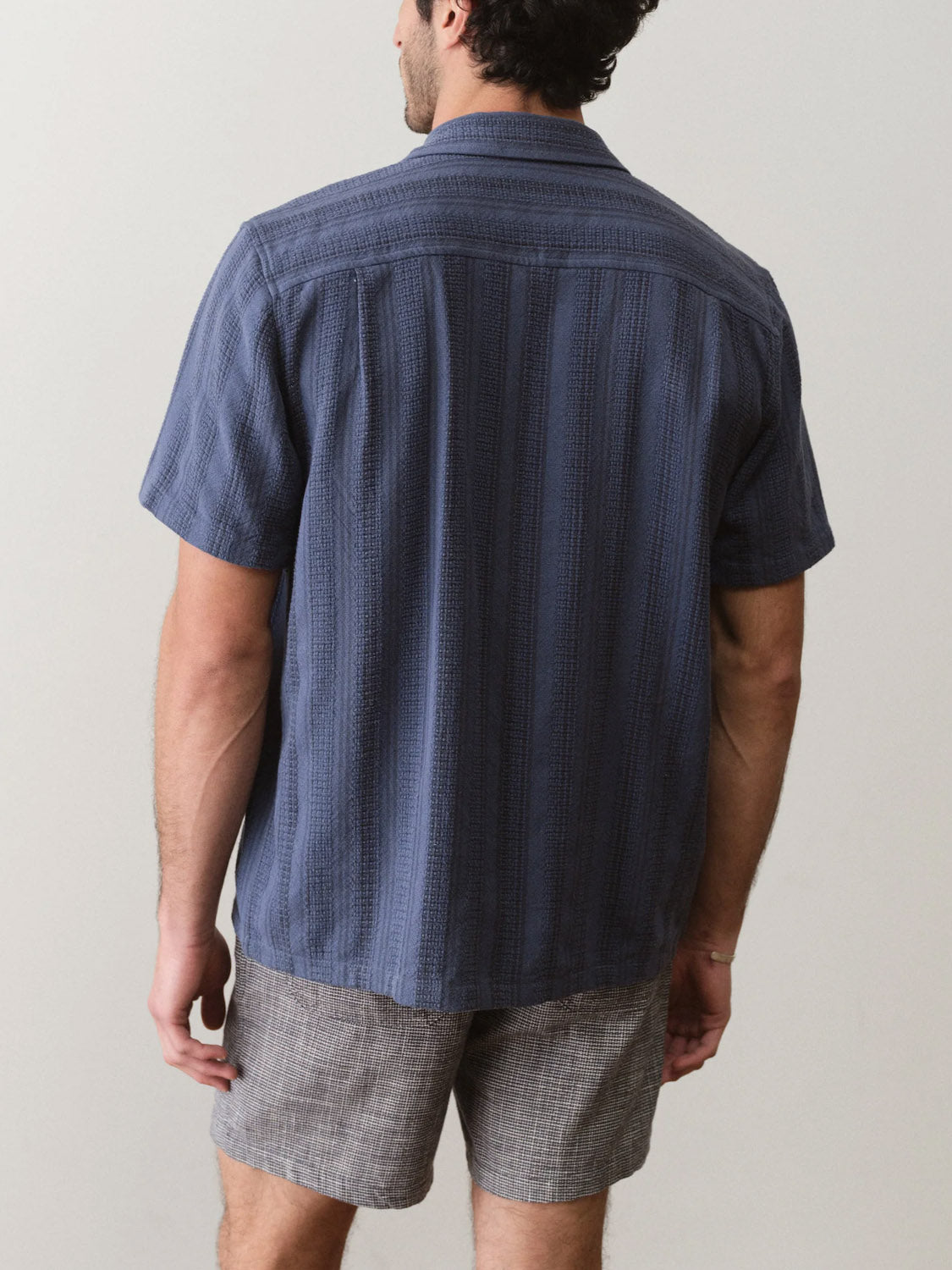 A man faces away from the camera, wearing the Marine Layer Dobby Resort Shirt in navy blue and gray shorts, set against a plain, light-colored background.