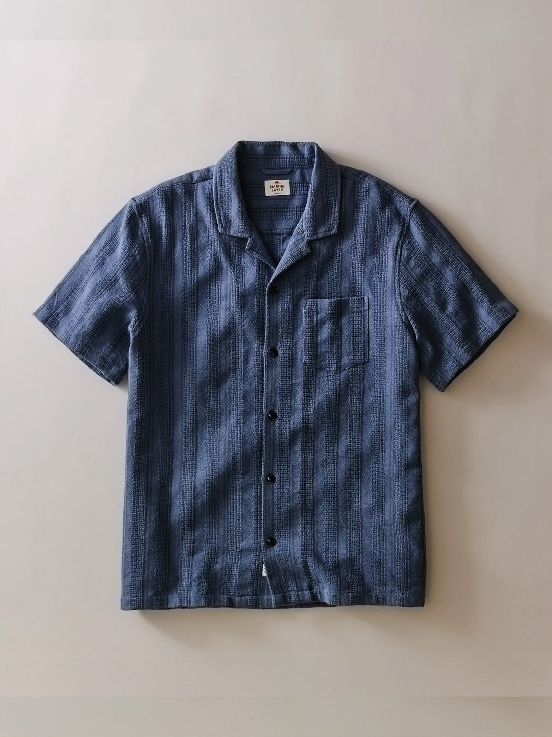 The Marine Layer Dobby Resort Shirt is a blue, short-sleeve button-up with a subtle striped texture, single chest pocket, and an inner collar label, shown laid flat on a light background.