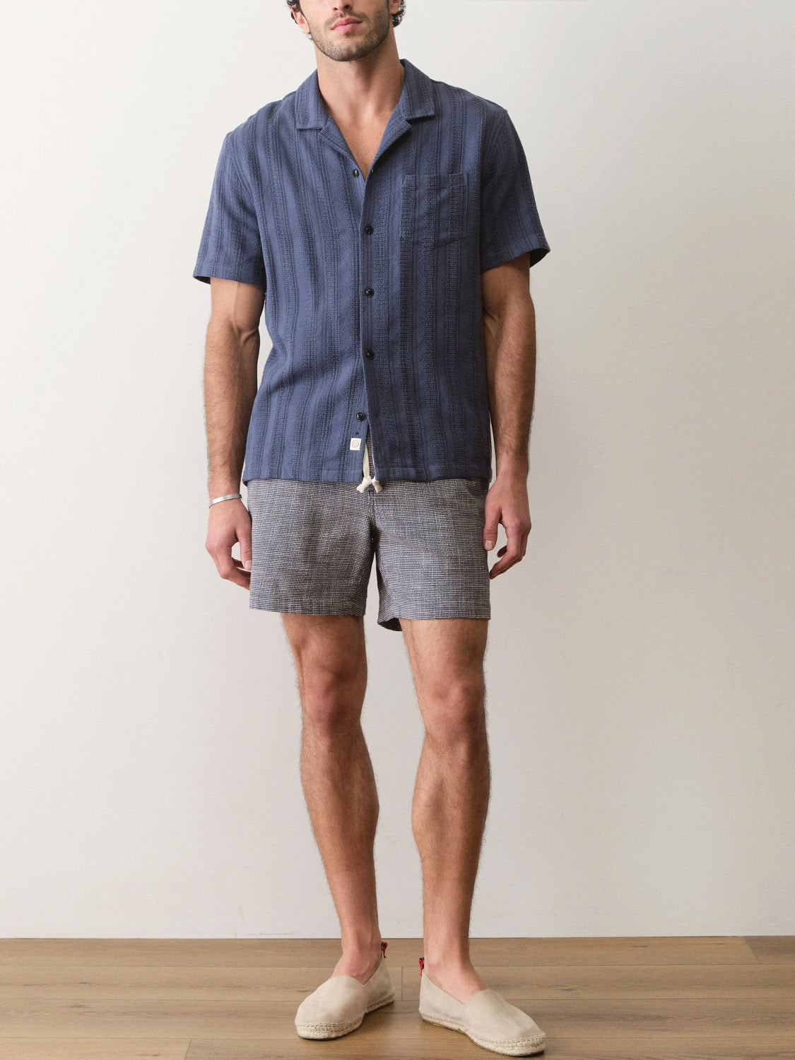 A man stands indoors against a plain wall, wearing the Marine Layer Dobby Resort Shirt in navy, gray shorts, and beige slip-on shoes. His face is partially out of the frame.