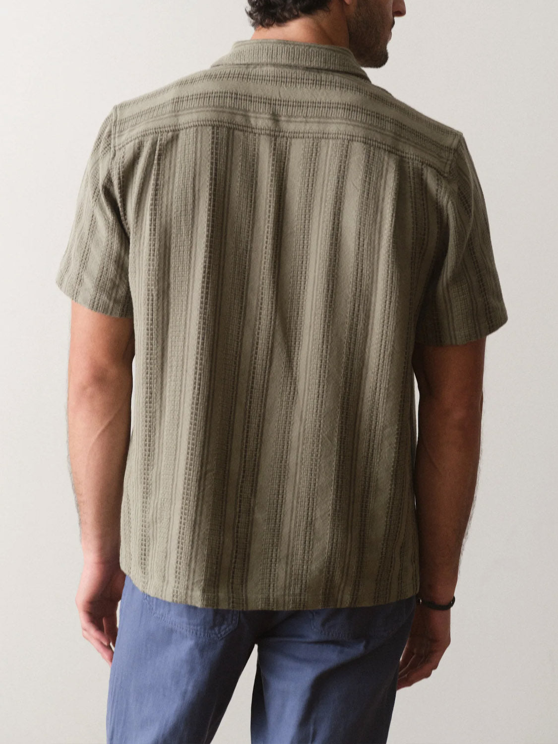 A man is seen from behind wearing the Marine Layer Dobby Resort Shirt in olive green with blue pants, standing against a plain light background.