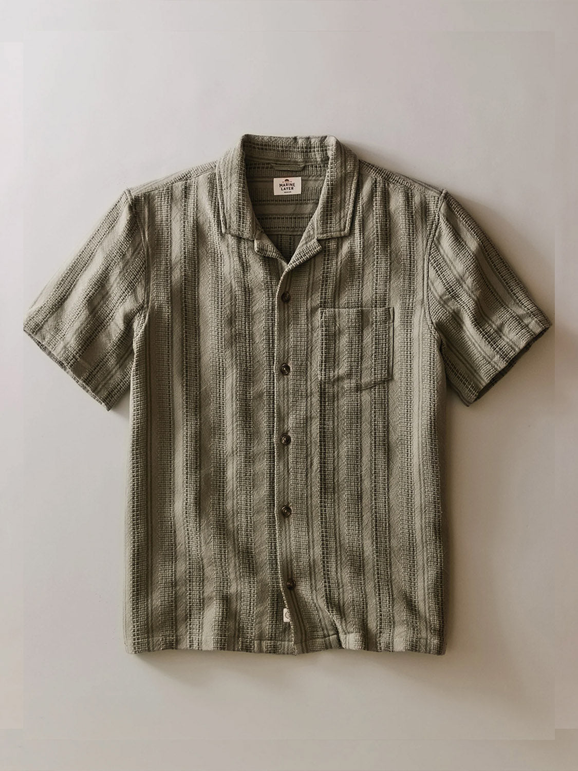 The Marine Layer Dobby Resort Shirt is a short-sleeve, button-up with a collar and chest pocket. Made from textured cotton, it features muted gray-green vertical stripes and is shown flat on a light background.