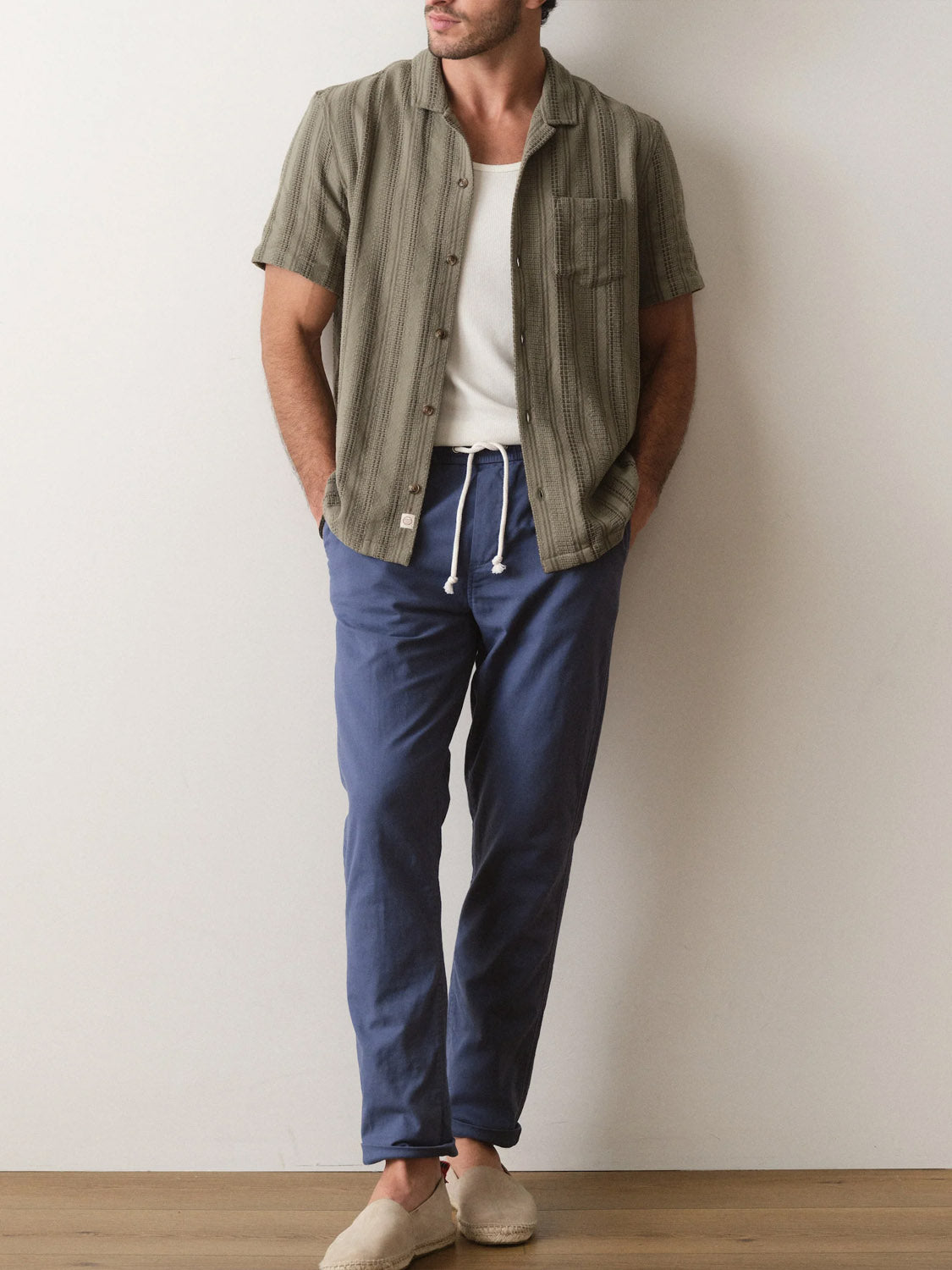 A man stands against a plain wall wearing Marine Layer's Dobby Resort Shirt over a white tee, blue drawstring pants, and beige slip-on shoes, hands in his pockets.
