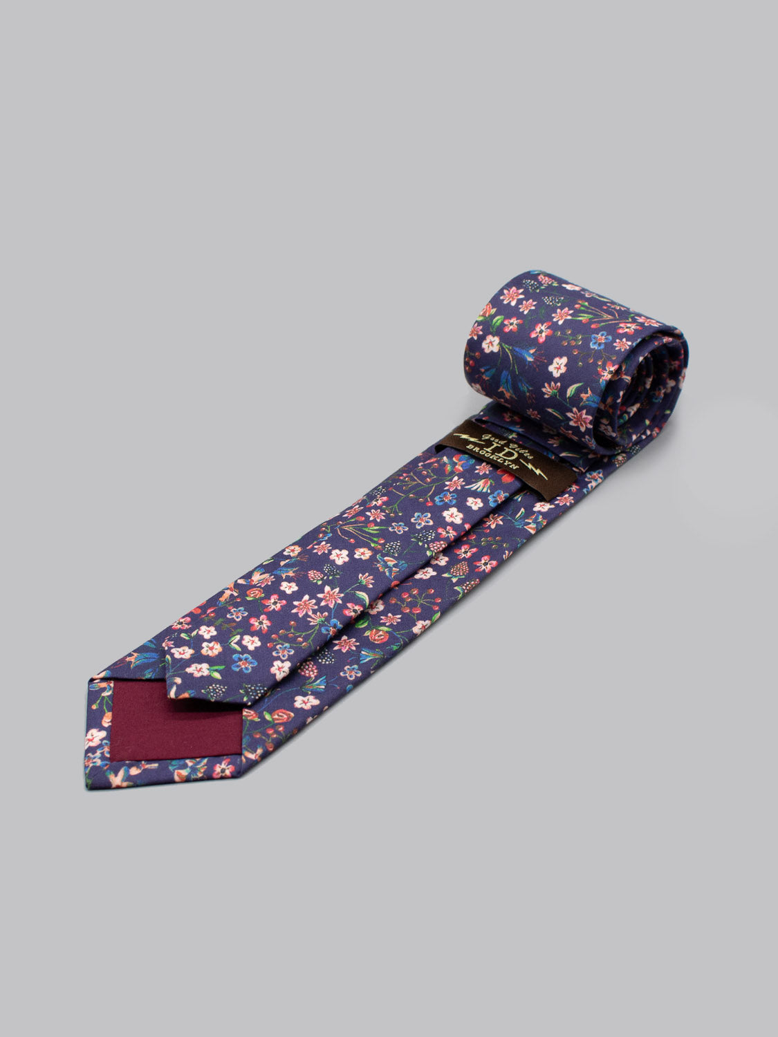 The Liberty of London Tana Lawn™ Donna Tie by ID Menswear features a colorful floral pattern on navy, handmade in Brooklyn from Tana Lawn cotton with a maroon inner tip accent, displayed on a light gray background.