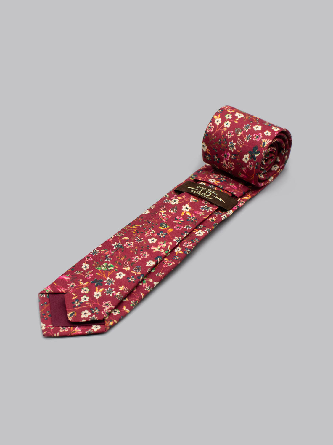 The Liberty of London Tana Lawn™ Donna Tie by ID Menswear, handmade in Brooklyn from fine cotton, displays a red floral pattern with yellow, white, and green accents, partially unrolled on a light gray background.