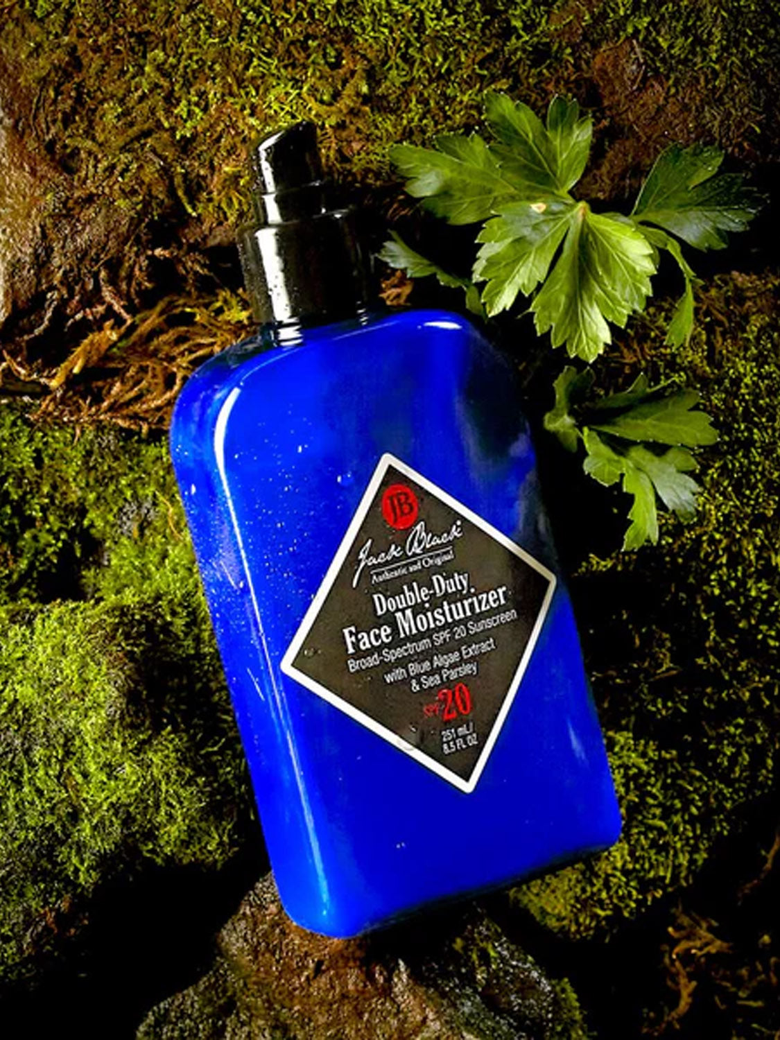 A blue bottle of Jack Black Double Duty Face Moisturizer SPF 20 sits on mossy rocks beside green leaves, emphasizing its broad-spectrum protection in a natural outdoor setting.