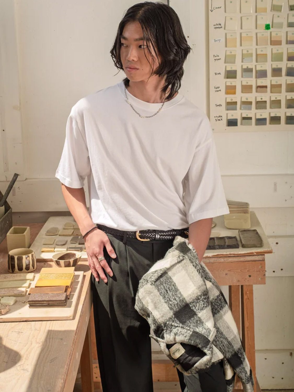 A person with shoulder-length hair stands in a studio wearing Kuwalla Double Pleated Pants, holding a black-and-white plaid jacket, and a wearing a white t-shirt.