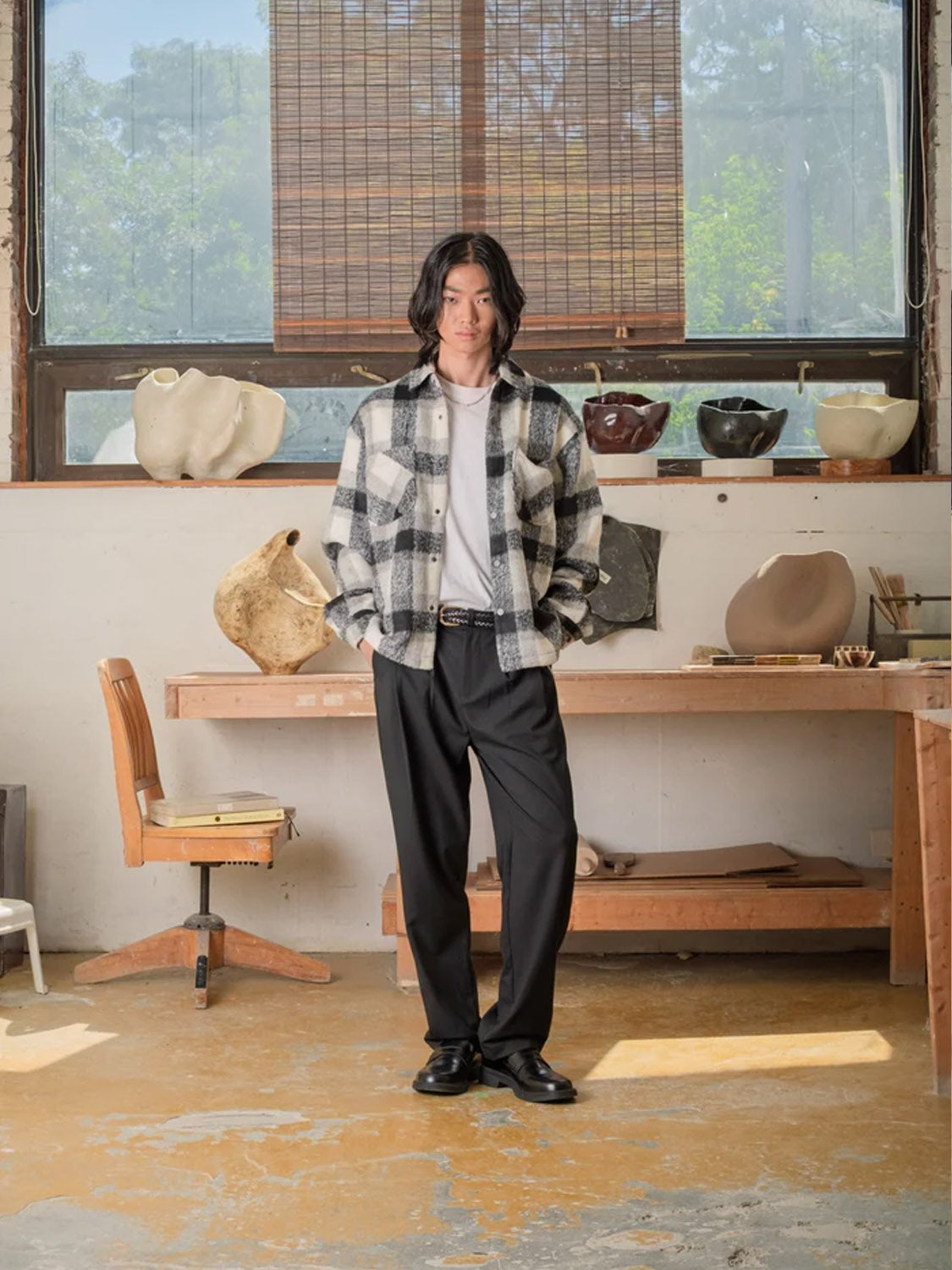 A person with shoulder-length hair stands in a studio wearing Kuwalla Double Pleated Pants, a black-and-white plaid jacket, and a white shirt. Shelves with ceramic pots, a large window, and an office chair are behind them.