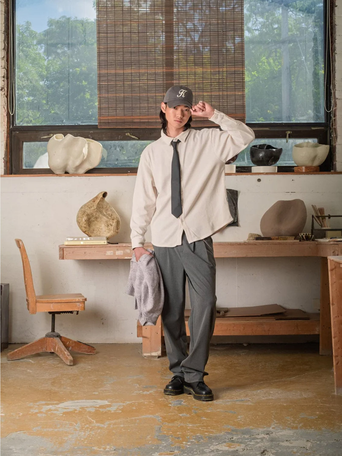 In an art studio, a person wears Kuwalla Double Pleated Pants with a light shirt, black tie, black shoes, and a cap. They hold a gray sweater, pottery and sculpting tools visible on tables behind them.