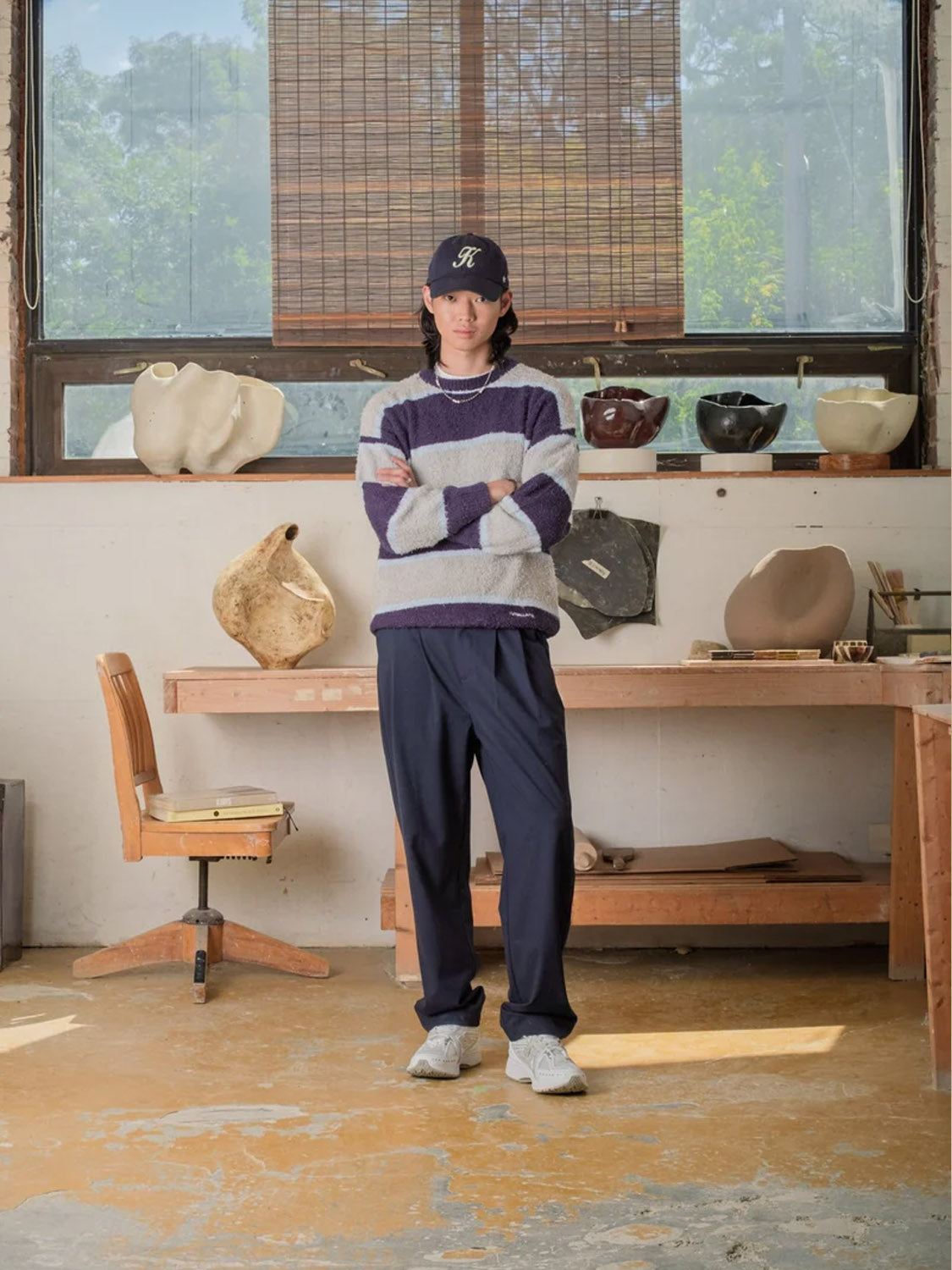 Wearing Kuwalla Double Pleated Pants, a person stands with arms crossed in a studio featuring pottery, a wooden chair, and a large window.