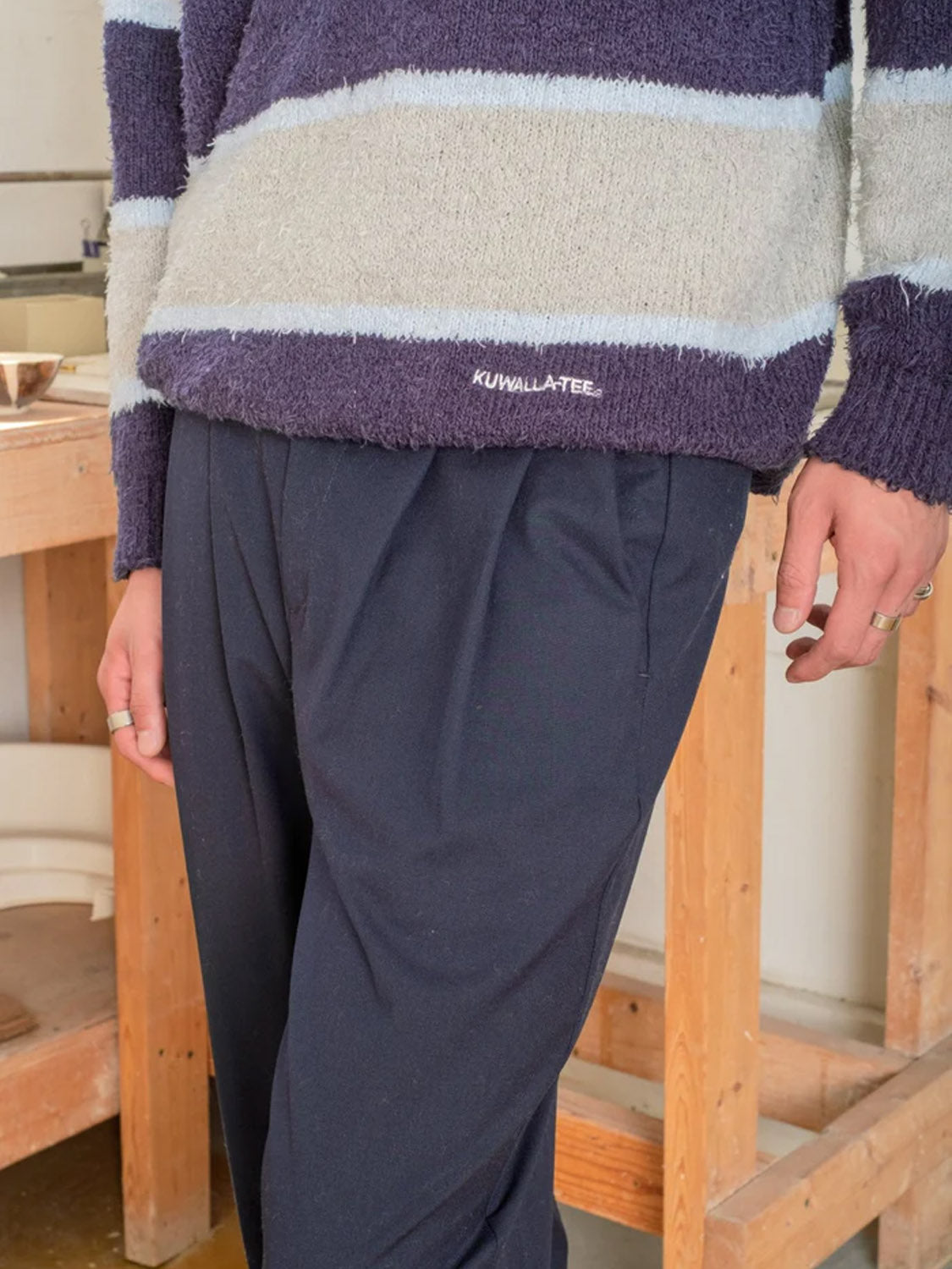 A person stands indoors near a wooden workbench wearing navy Double Pleated Pants from Kuwalla and a fuzzy striped sweater. The upper part of their body is partially cropped out of the image.