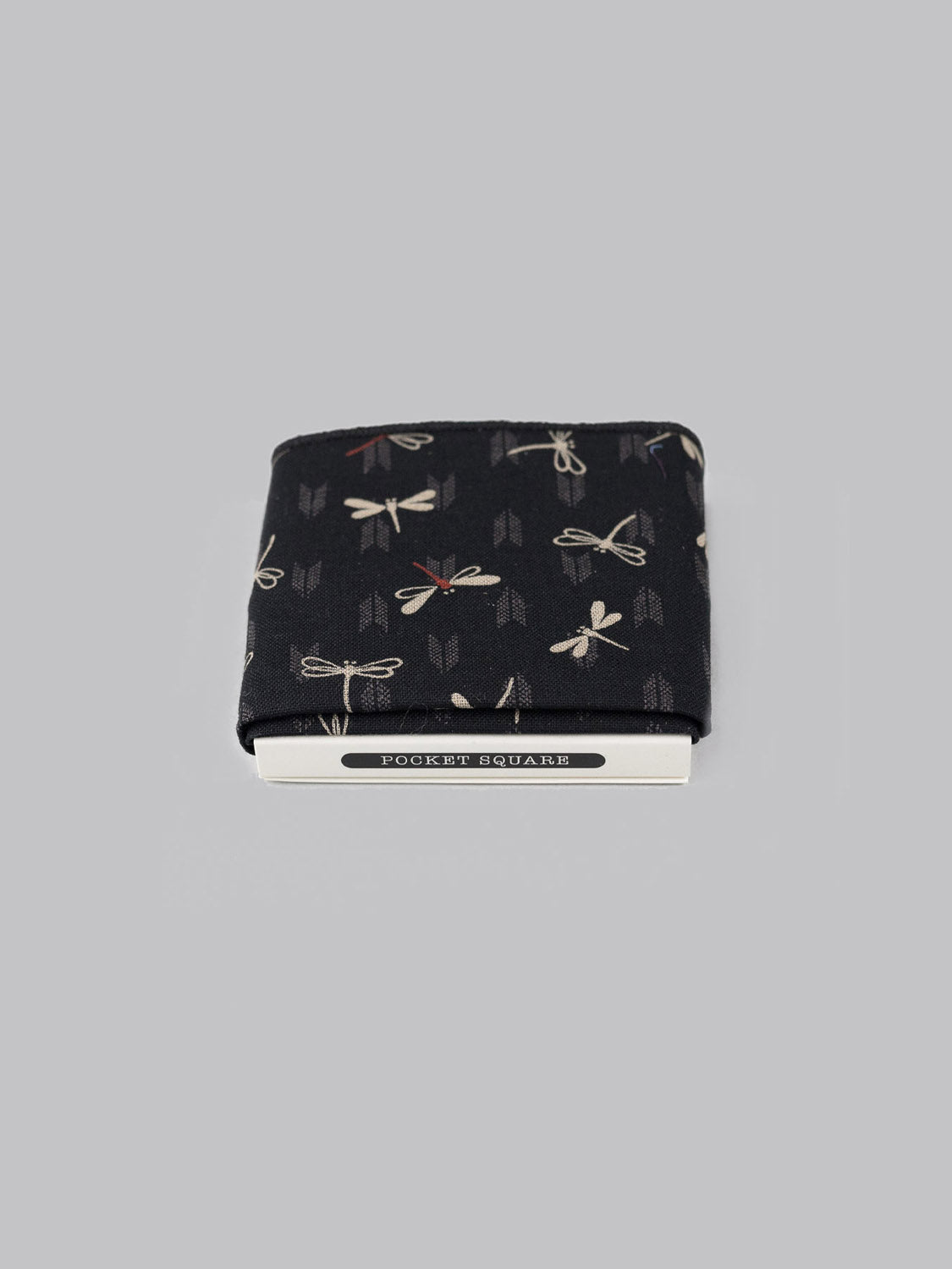 The Dragonfly Pocket Square by ID Menswear, a handmade black cotton accessory with a dragonfly pattern, is neatly folded and partially tucked into white POCKET SQUARE packaging, set against a plain gray background.