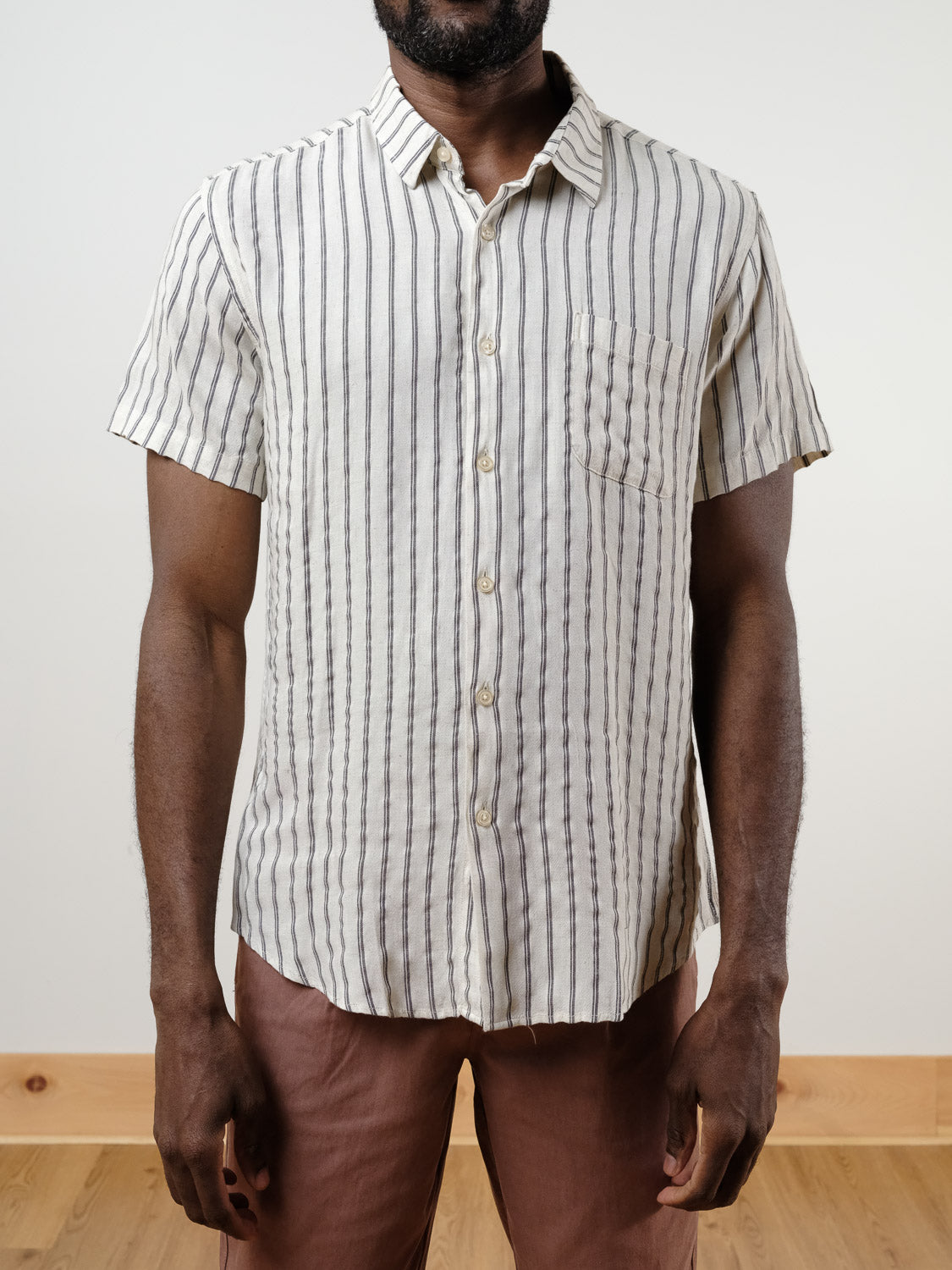 A man wears the Drifter Linen Rayon Blend Shirt by ID Menswear, a lightweight short-sleeved top with vertical black and white stripes, paired with brown pants, standing indoors against a plain background.