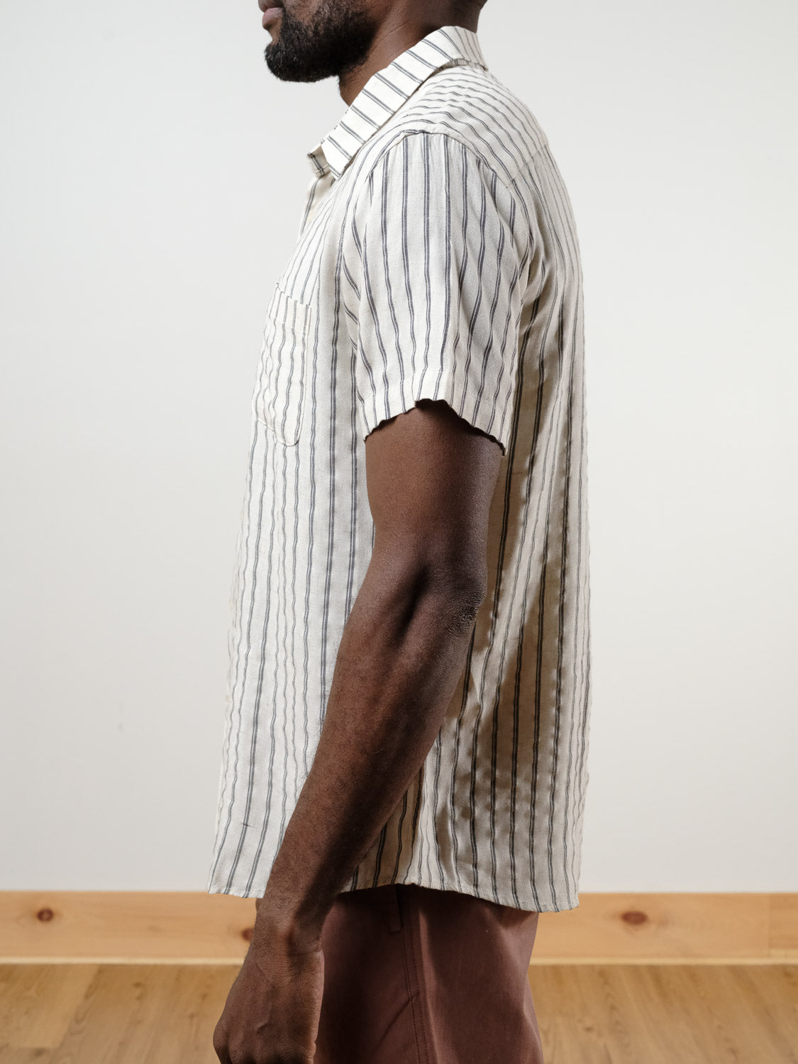 A man stands in profile wearing the Drifter Linen Rayon Blend Shirt by ID Menswear—a beige, short-sleeved button-up with vertical black stripes. The background is plain with light walls and a wooden floor.