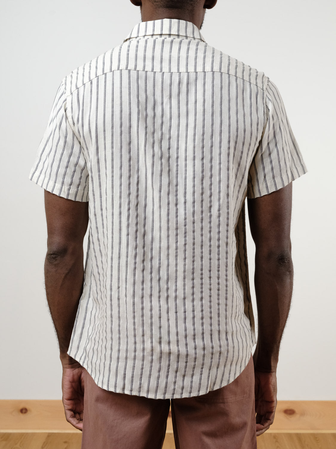A person shown from the back against a plain background is wearing the Drifter Linen Rayon Blend Shirt by ID Menswear, a lightweight short-sleeved woven stripe shirt, paired with light brown pants.