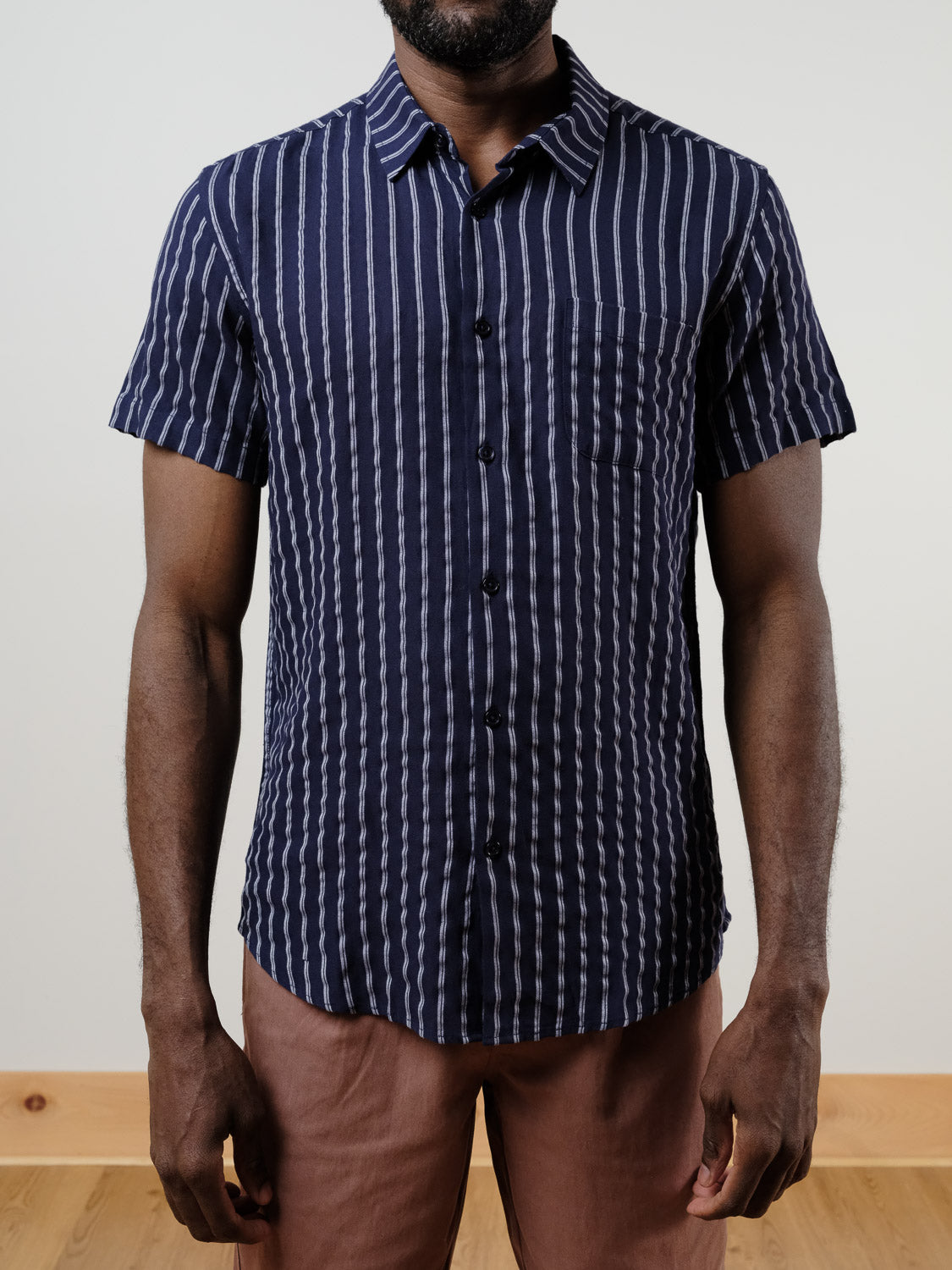 A man wearing the ID Menswear Drifter Linen Rayon Blend Shirt, a lightweight short-sleeved navy shirt with vertical white stripes, and light brown pants stands against a plain, light-colored background.