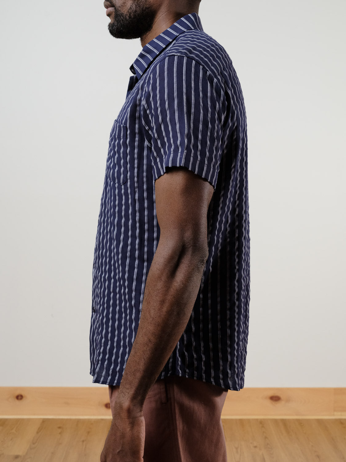 A man stands in profile wearing the Drifter Linen Rayon Blend Shirt by ID Menswear—a lightweight, short-sleeved navy blue shirt with vertical white stripes—paired with brown pants. The background features light walls and a wooden floor.